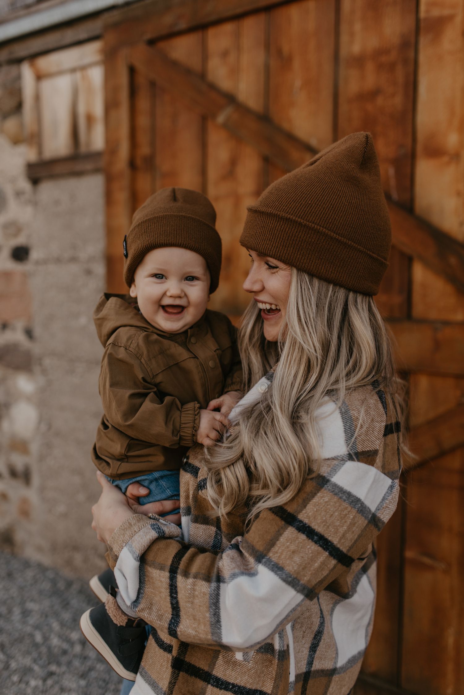 A smiling mother and baby wearing matching brown beanies and warm outfits, sharing a joyful moment outdoors near a wooden barn.