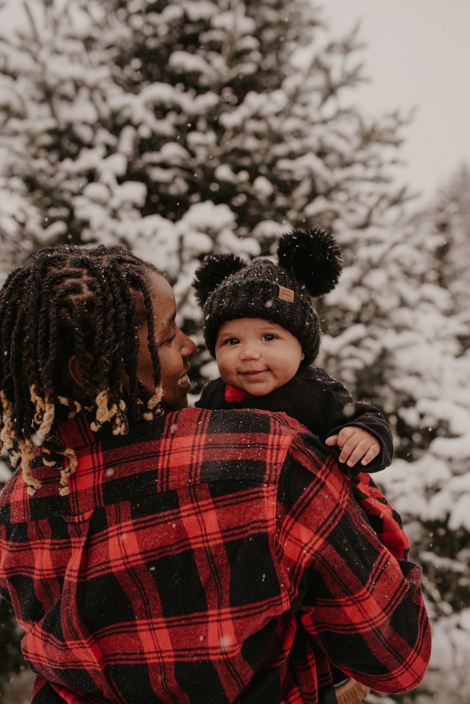 Parent in red plaid shirt holds smiling baby in black pom-pom hat outdoors among snow-covered evergreen trees.