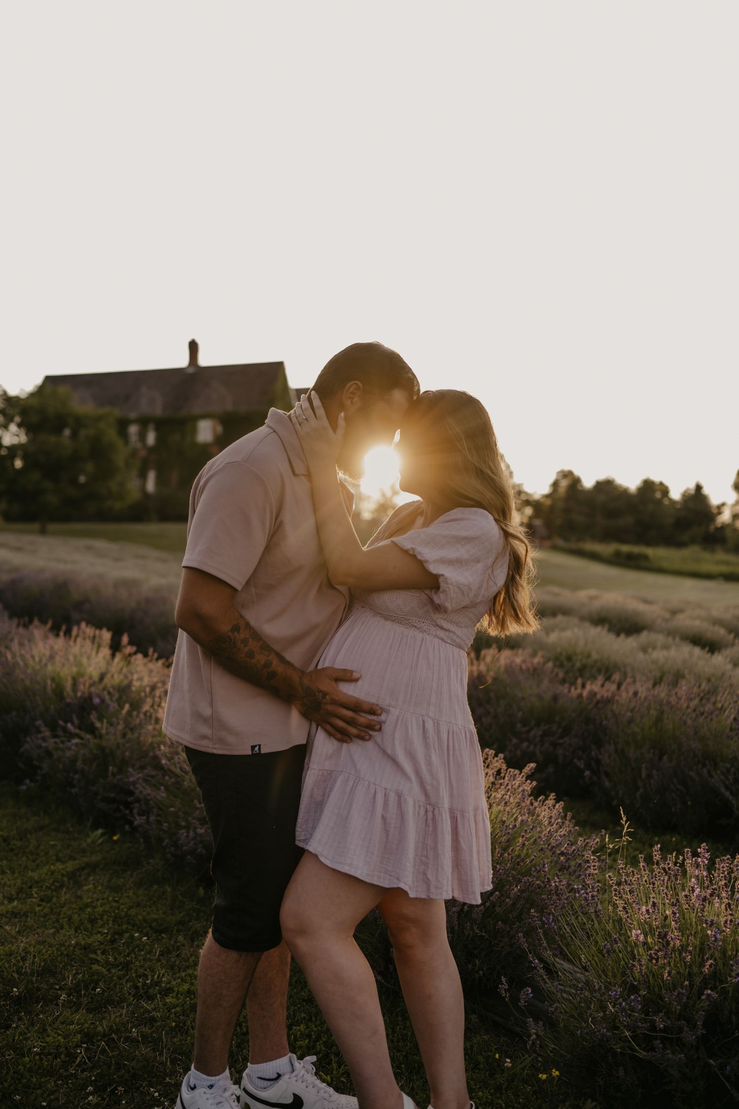 Couple sharing a golden hour kiss in a lavender field, sun glowing between them, woman in pink dress.