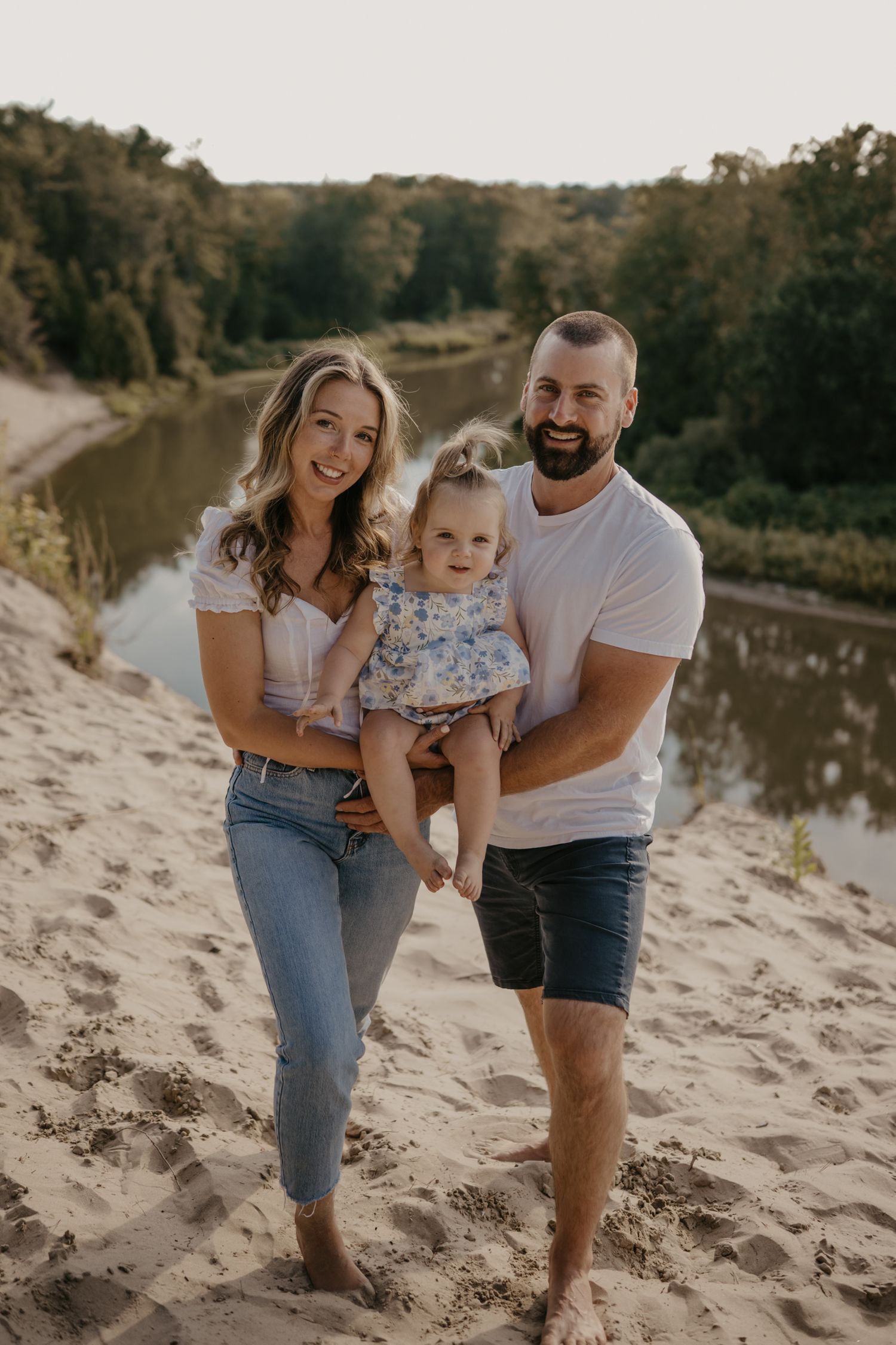 Happy family of three posing on sandy beach dunes with river and trees in background on sunny day.
