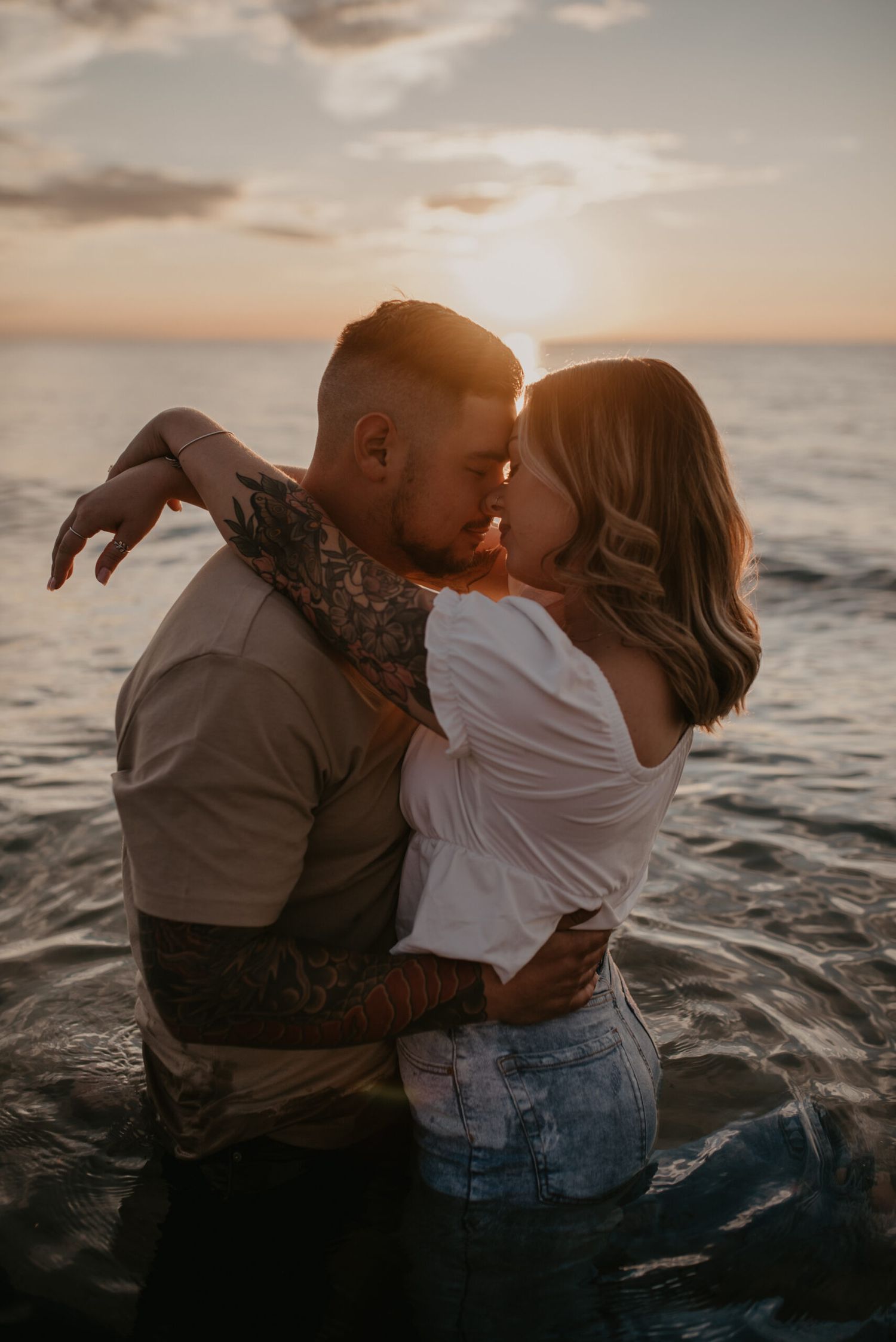 Couple shares passionate kiss standing in ocean waves during golden sunset.