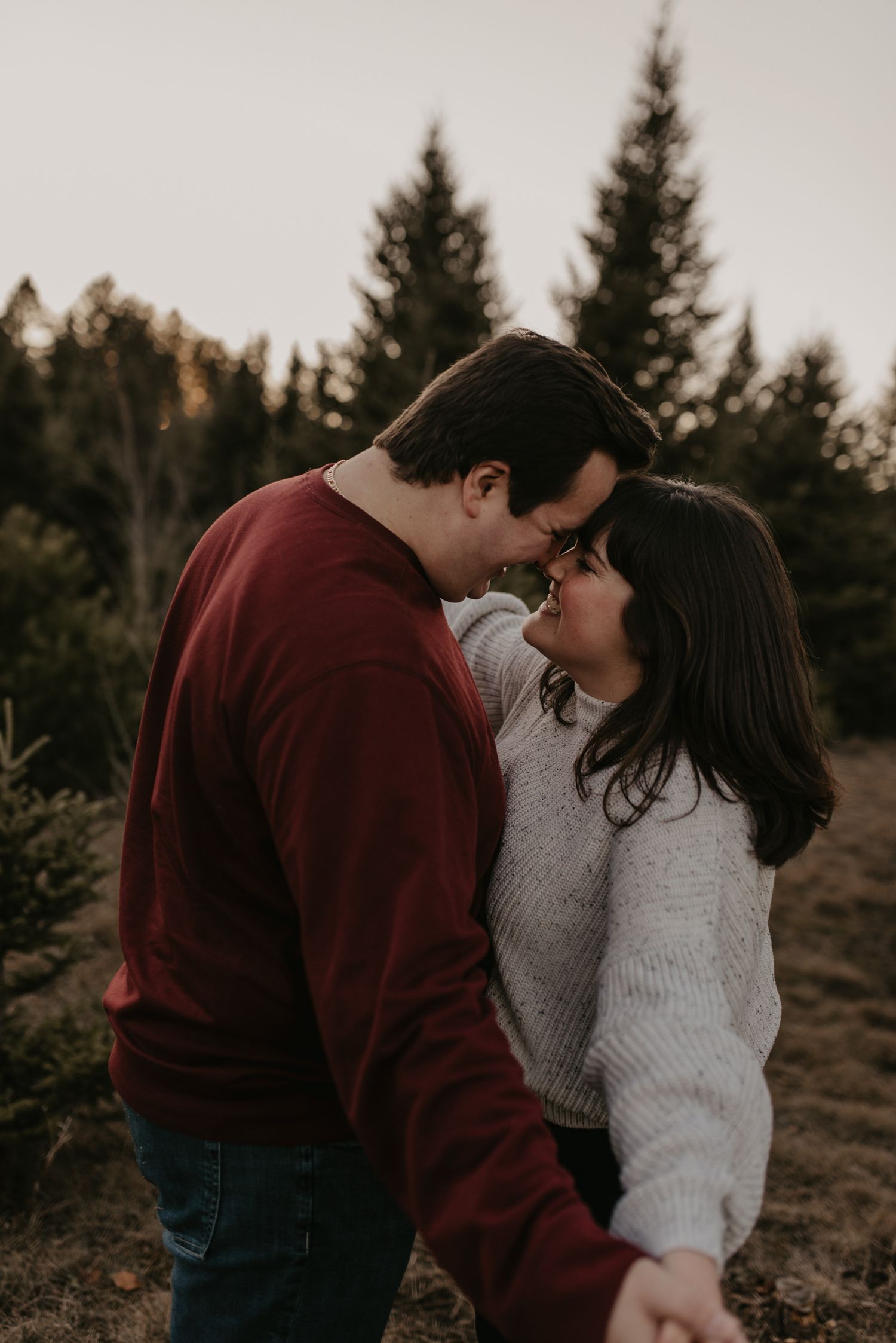 Young couple touching foreheads and smiling tenderly in evergreen forest at dusk.
