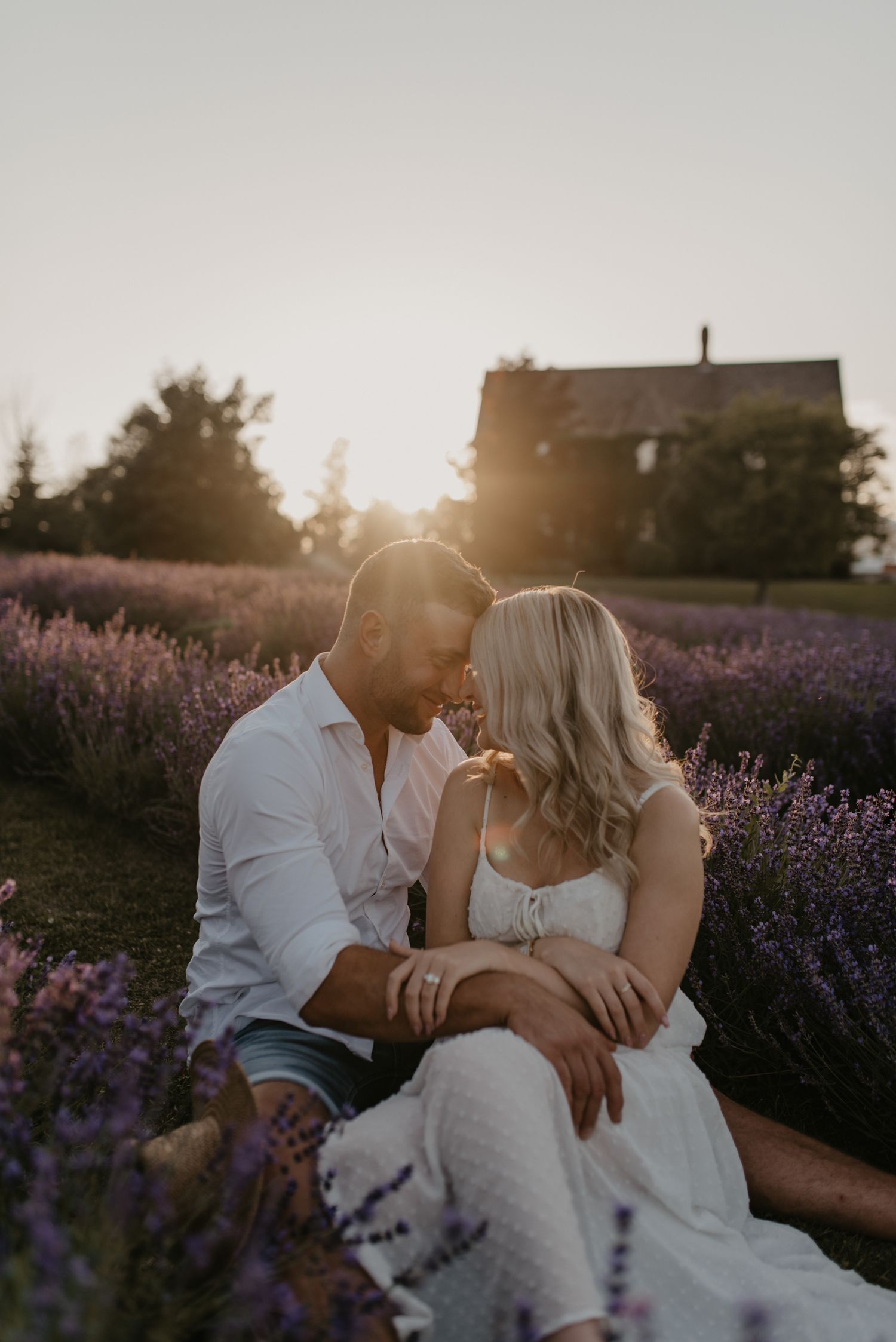 Couple sitting close together in blooming lavender field with warm golden sunset light behind them.