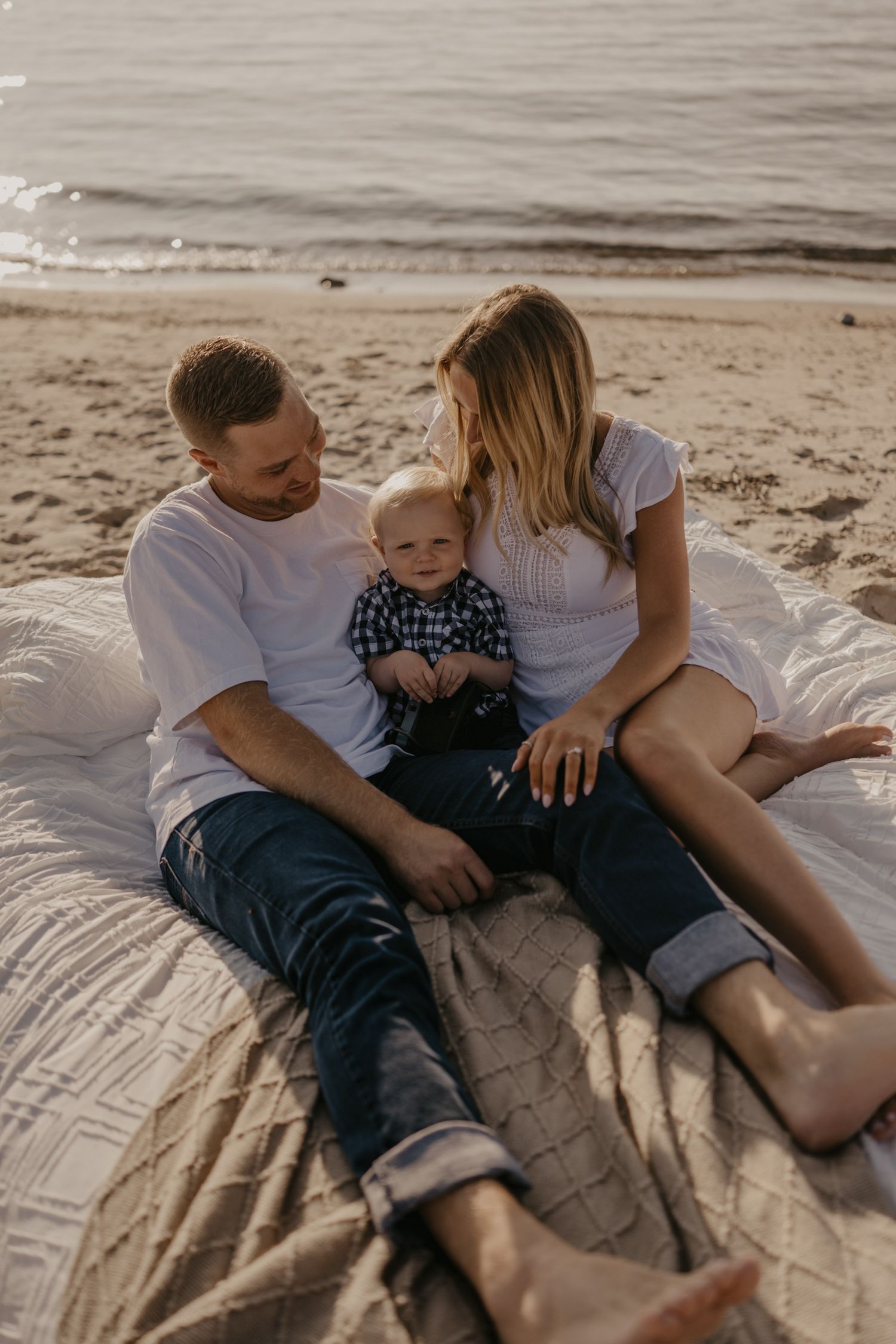Young family with baby boy sitting together on beach blanket near ocean at sunset.