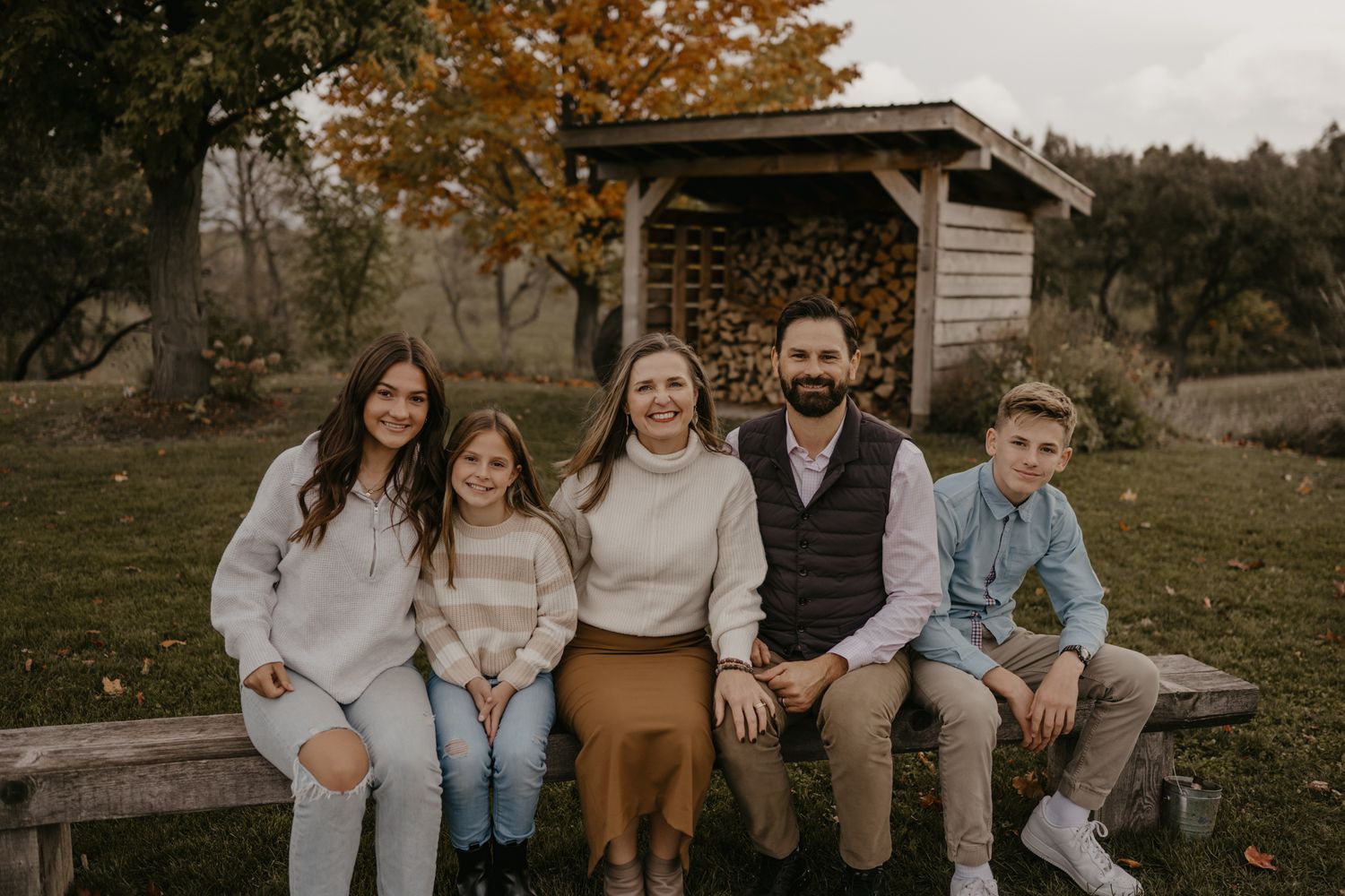 Family of five smiling together on a wooden bench outdoors in autumn, with colorful fall foliage and a firewood shed behind them.