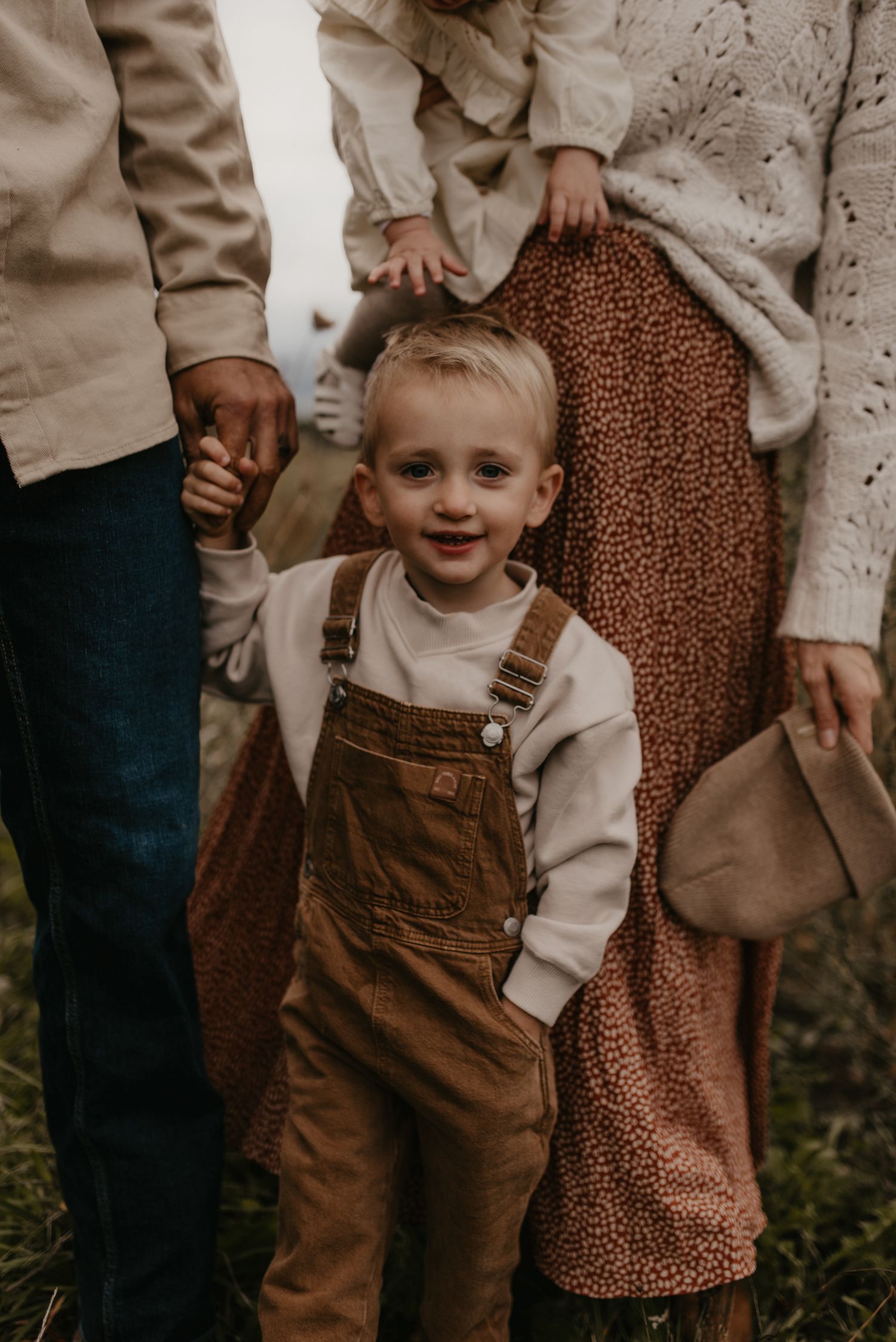 Adorable blonde toddler in brown overalls smiling outdoors during a fall family portrait session.