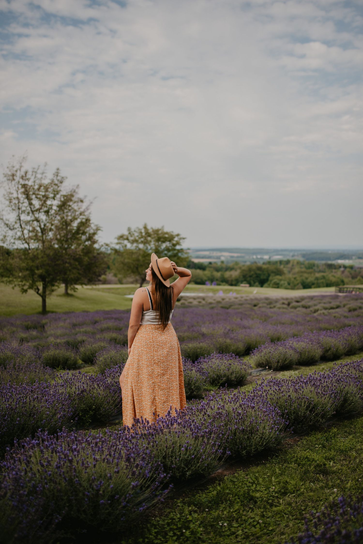 Woman in orange maxi skirt and hat standing among rows of purple lavender in a blooming field.