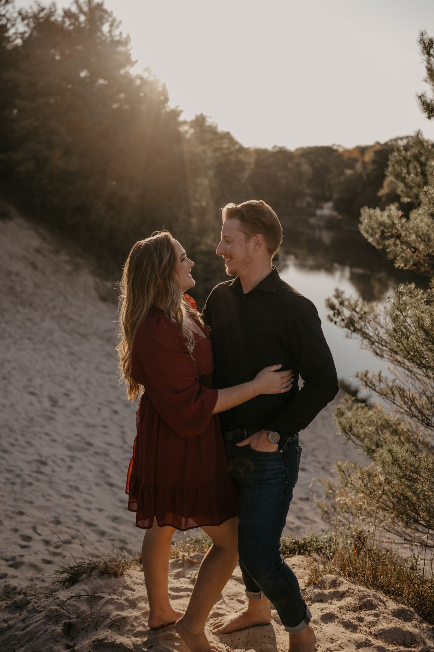 Couple embracing and smiling at each other on sandy dunes with sunlit river and trees backdrop.