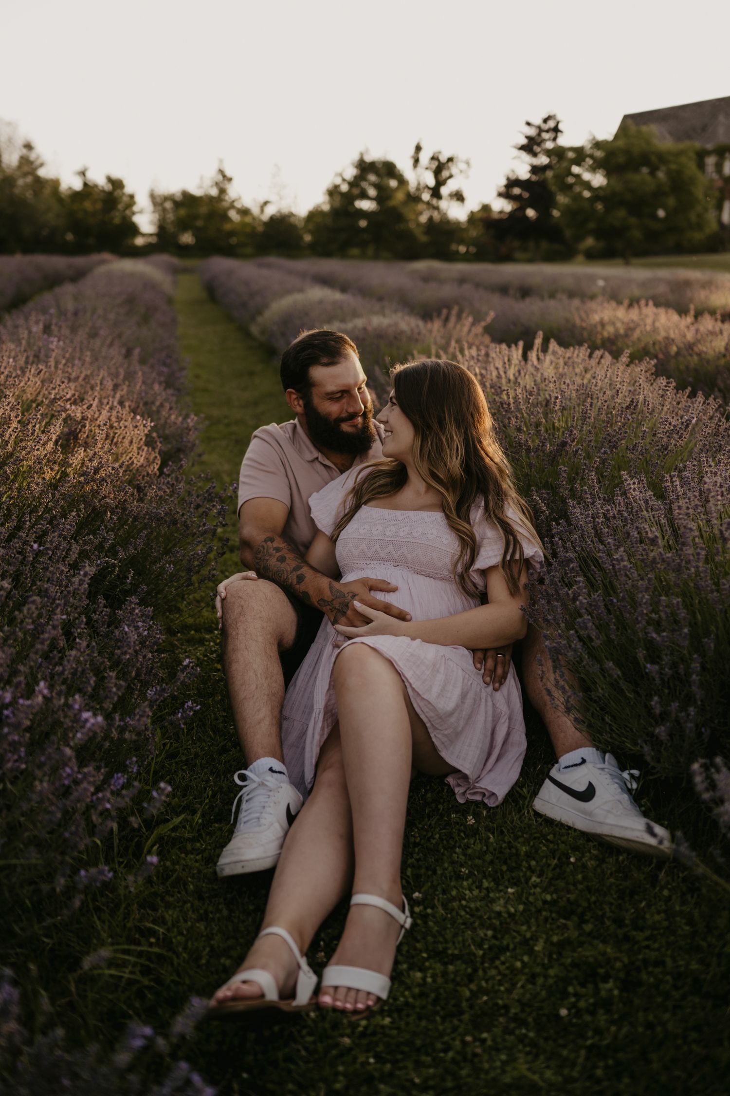 Couple sitting together in lavender field at sunset, man embracing pregnant woman in pink dress.