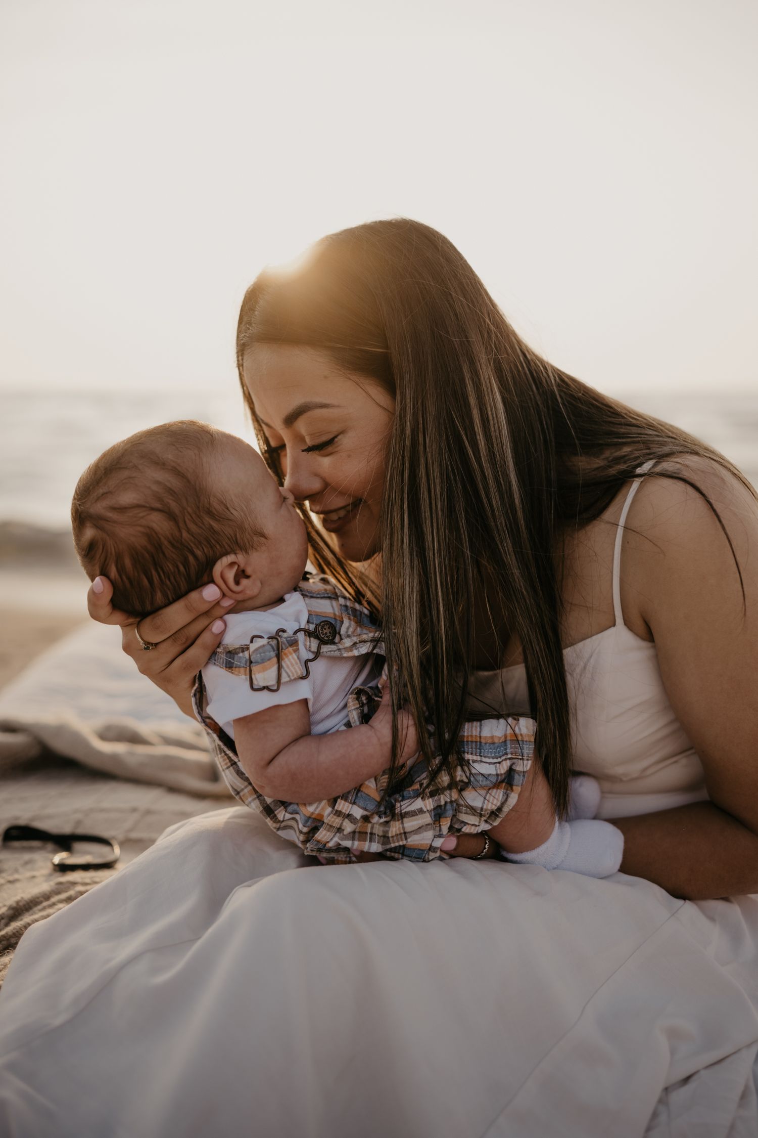 Mother tenderly kissing newborn baby on beach during golden hour sunset.