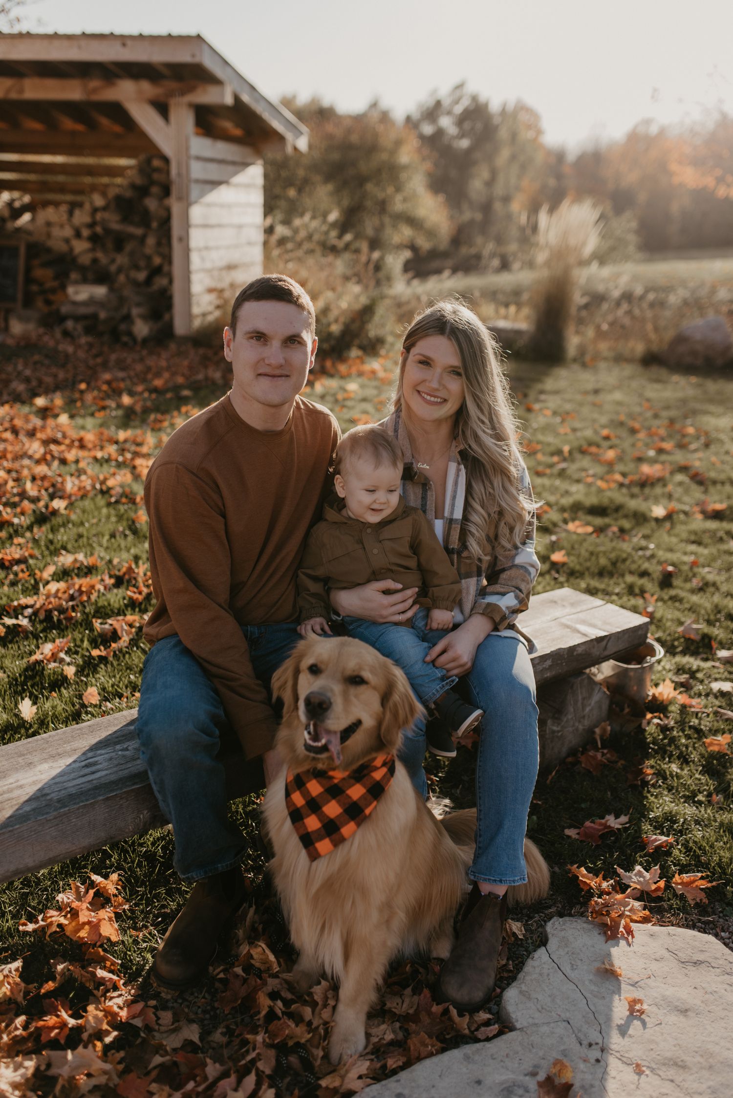 Family portrait with couple, baby, and golden retriever in buffalo plaid bandana, sitting outdoors among fall foliage.