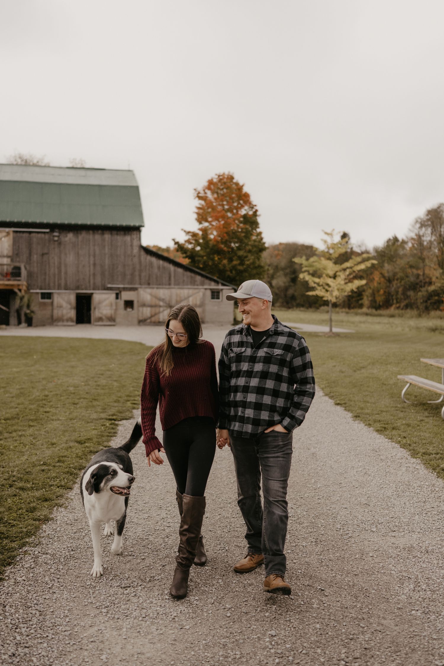 Couple walking dog on gravel path near rustic barn in autumn countryside setting.