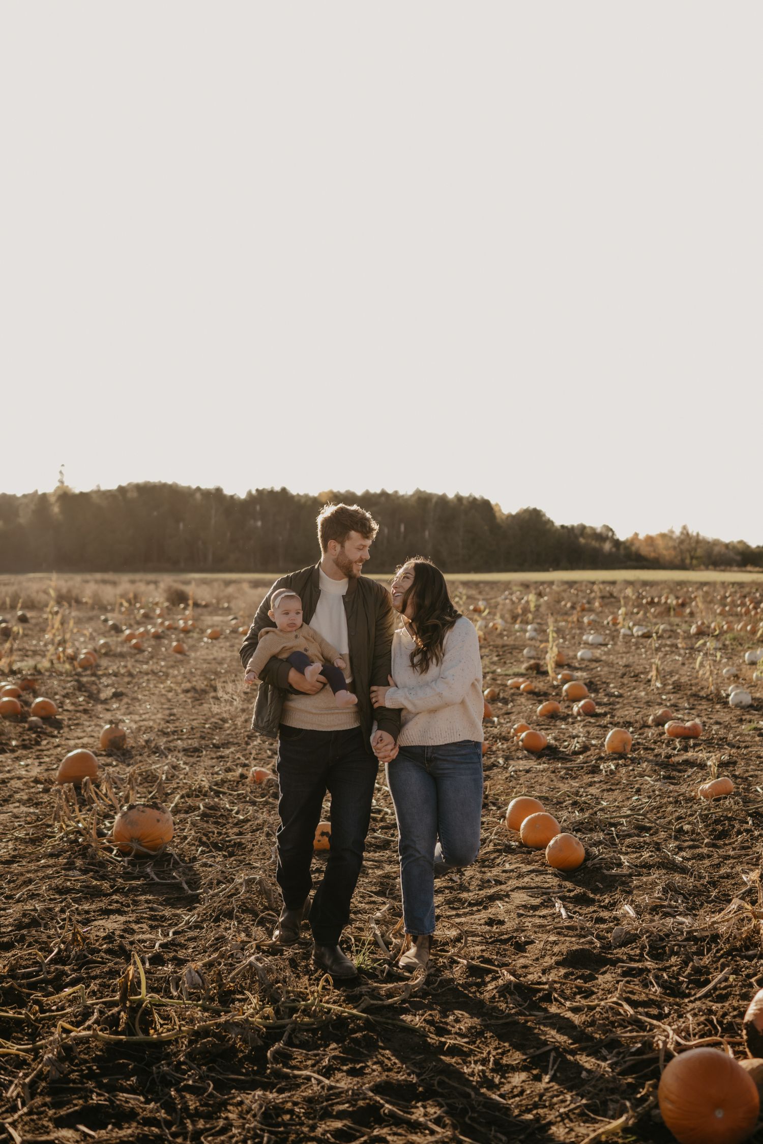 A young family walks through a pumpkin patch in autumn, with the man holding a baby and the woman smiling beside him.