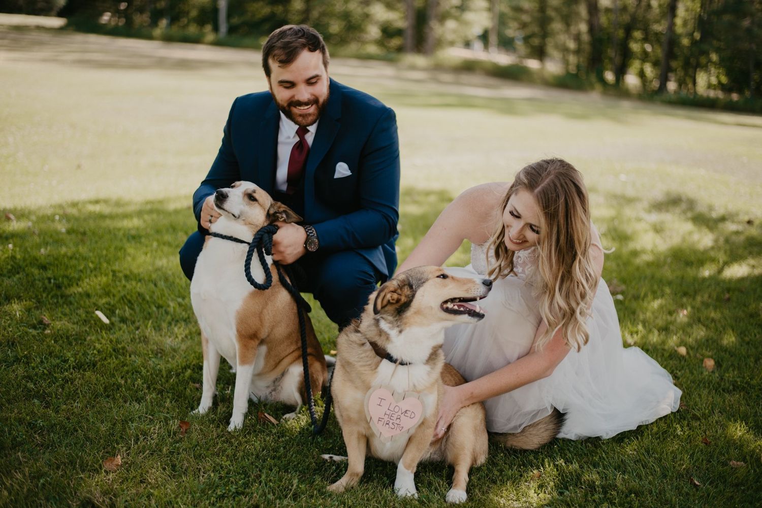 Smiling bride and groom crouching on grass with two dogs on leashes at outdoor wedding.