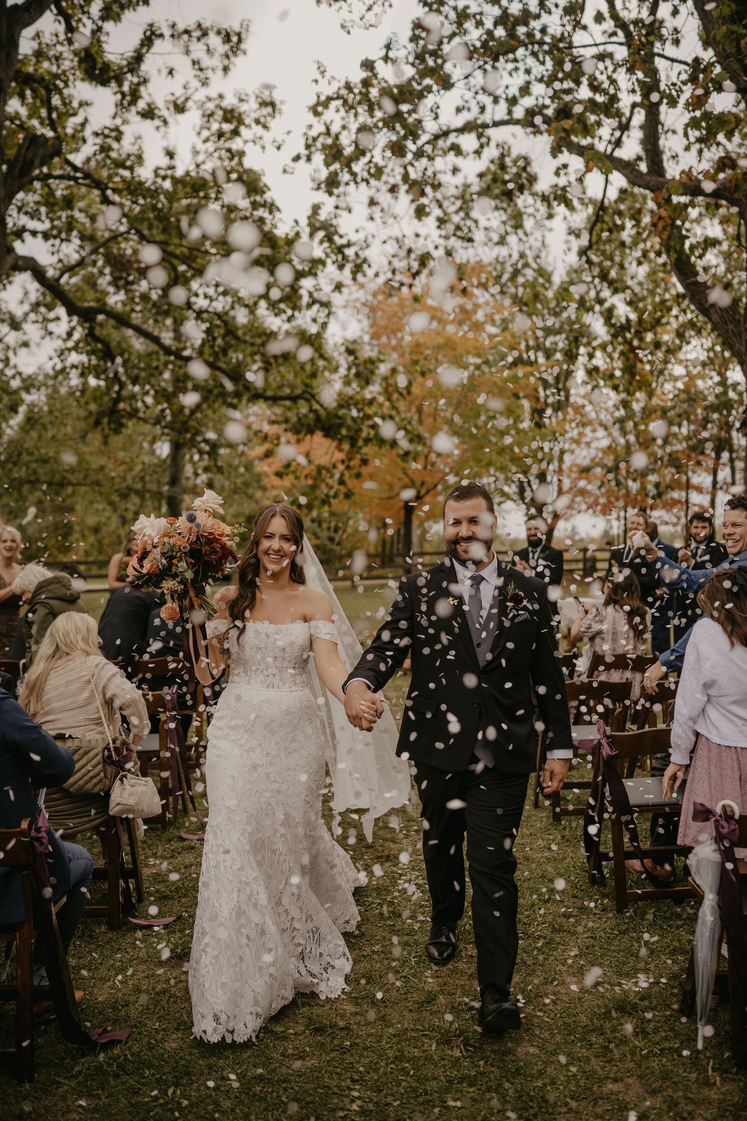Joyful newlyweds walking down the aisle through a shower of white confetti outdoors.