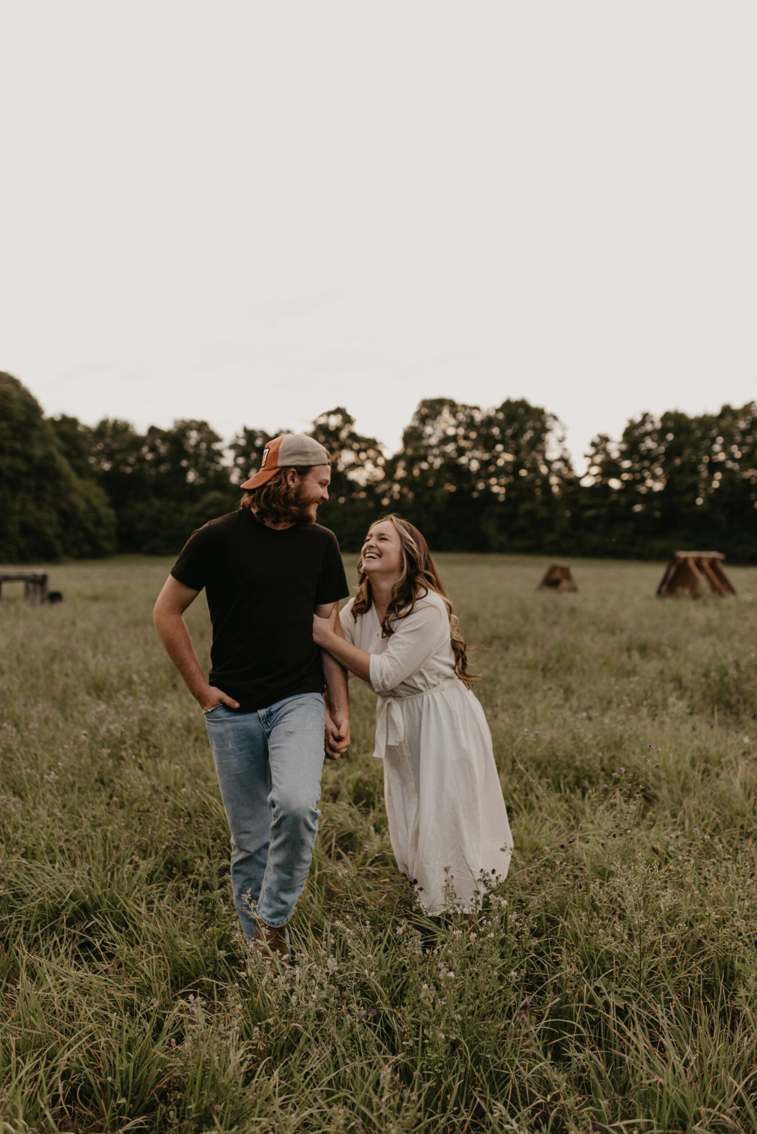 Couple laughing and holding hands while walking through a lush green meadow at golden hour.