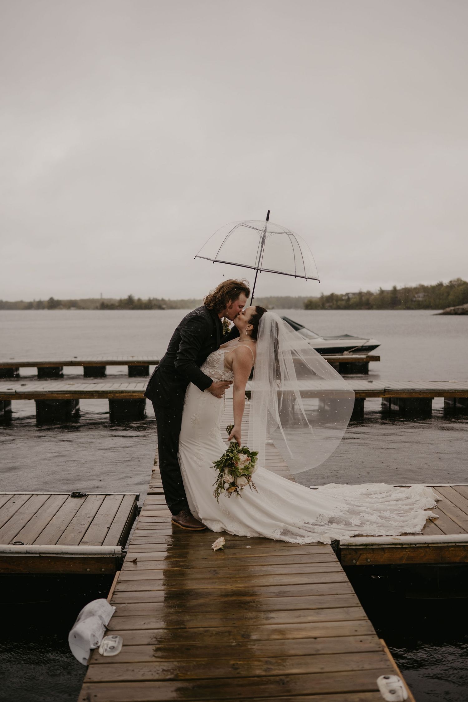Bride and groom kissing on a rainy dock under a clear umbrella by the lake.