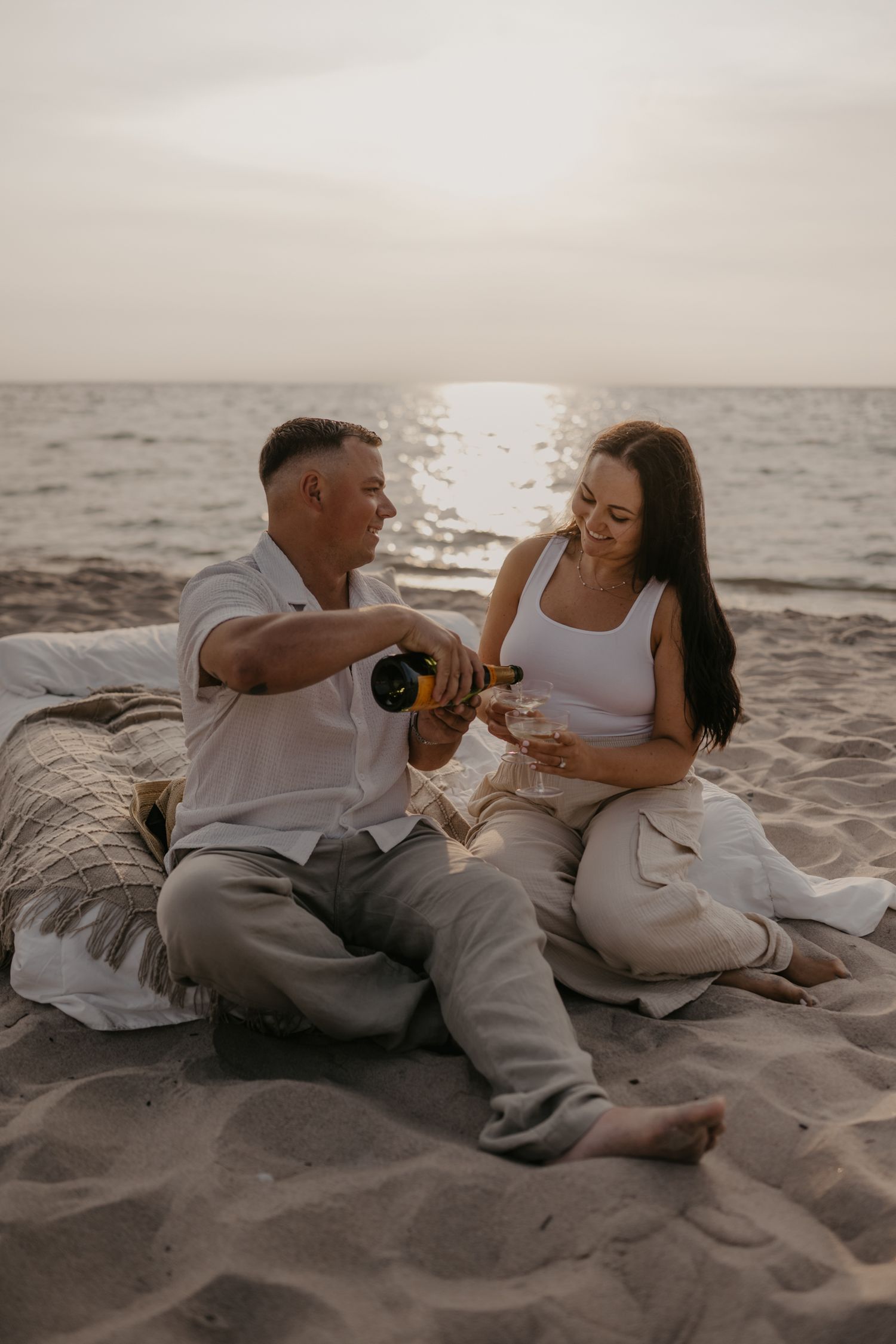 Couple enjoying champagne on sandy beach at sunset, man pouring wine for smiling woman.