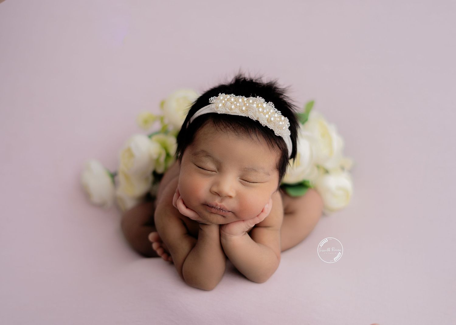 Smiling newborn baby girl in froggy pose with pearl headband surrounded by white flowers on lavender backdrop.