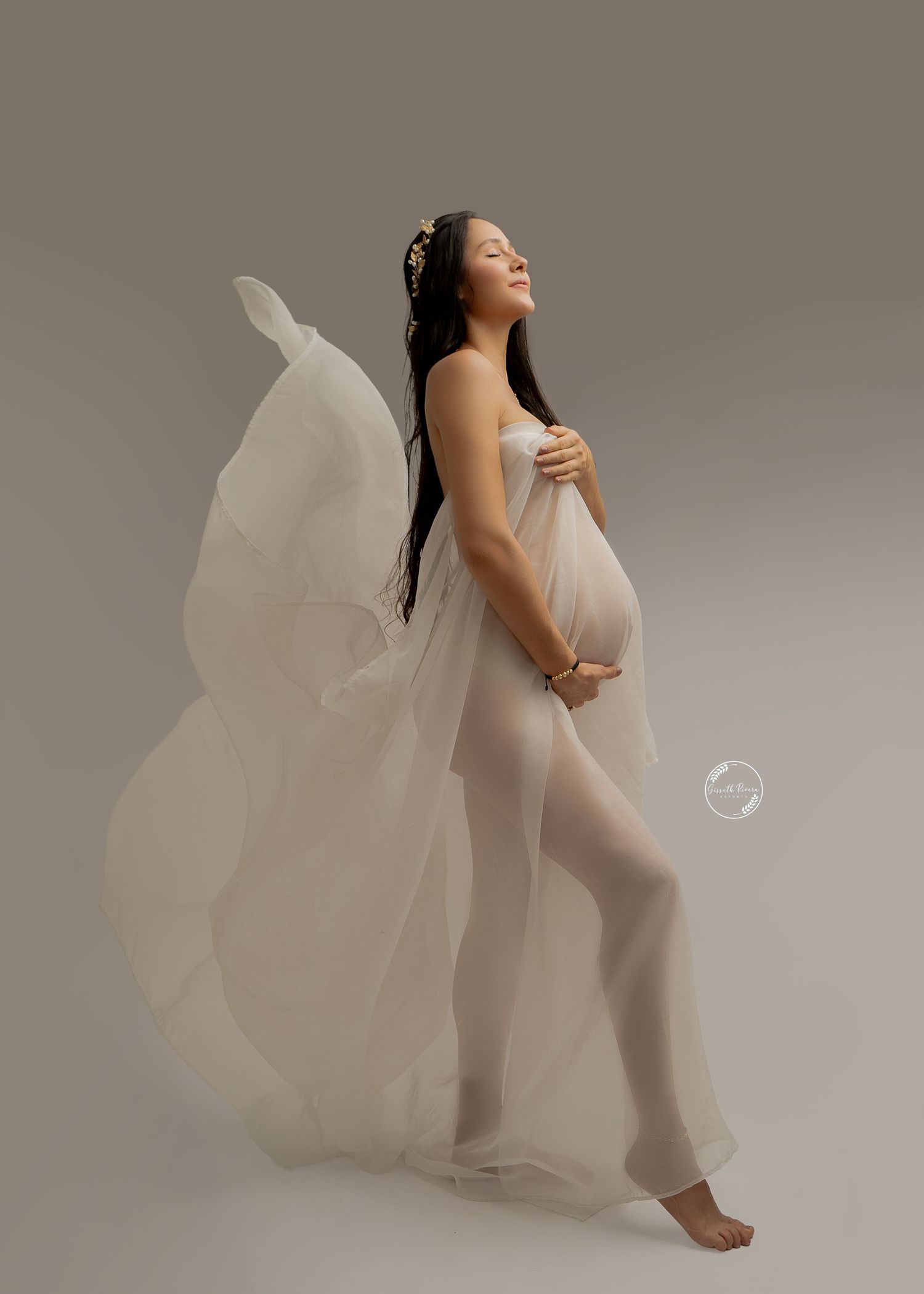 Expecting mother in flowing white sheer fabric poses gracefully in artistic maternity studio photo.