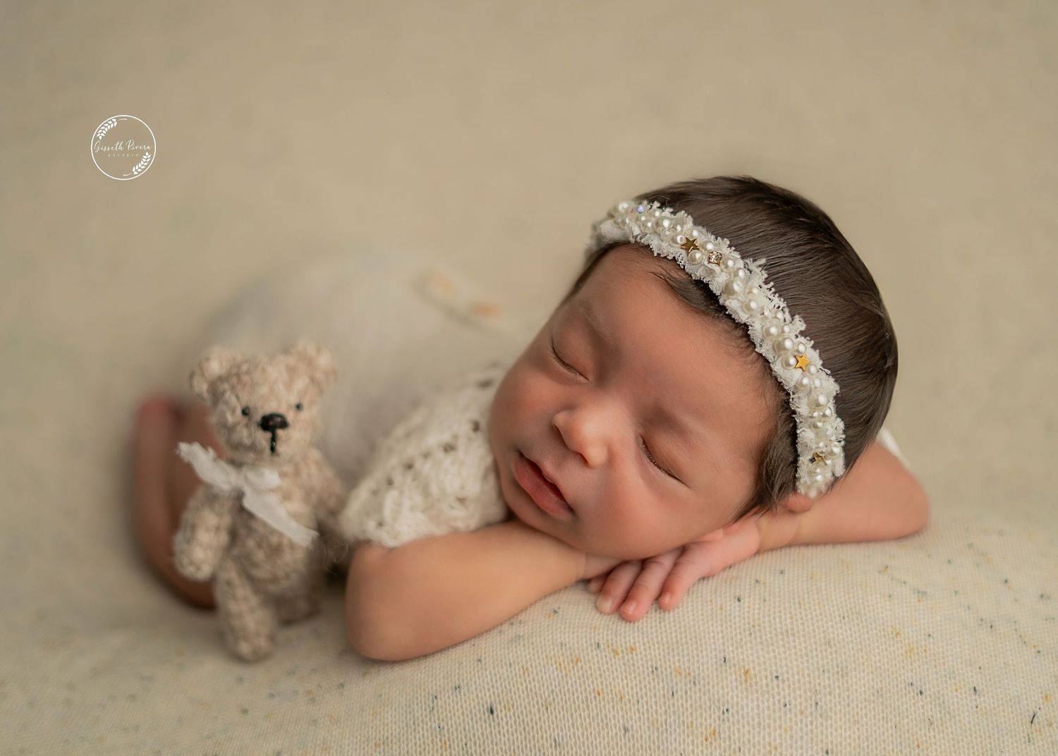Peaceful newborn baby sleeping with teddy bear wearing floral headband on beige backdrop.