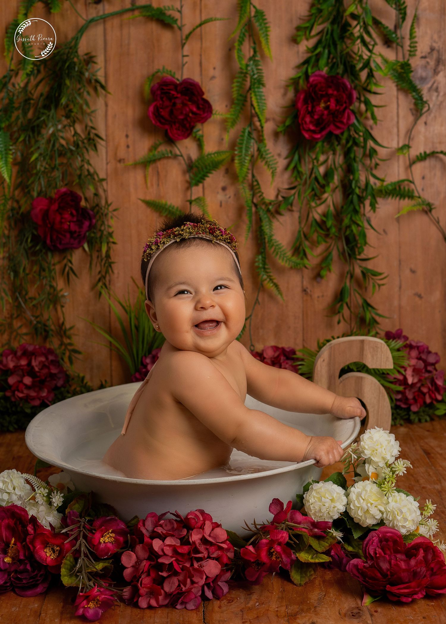 Happy baby laughing in a white basin surrounded by red and white flowers against a rustic wooden backdrop.