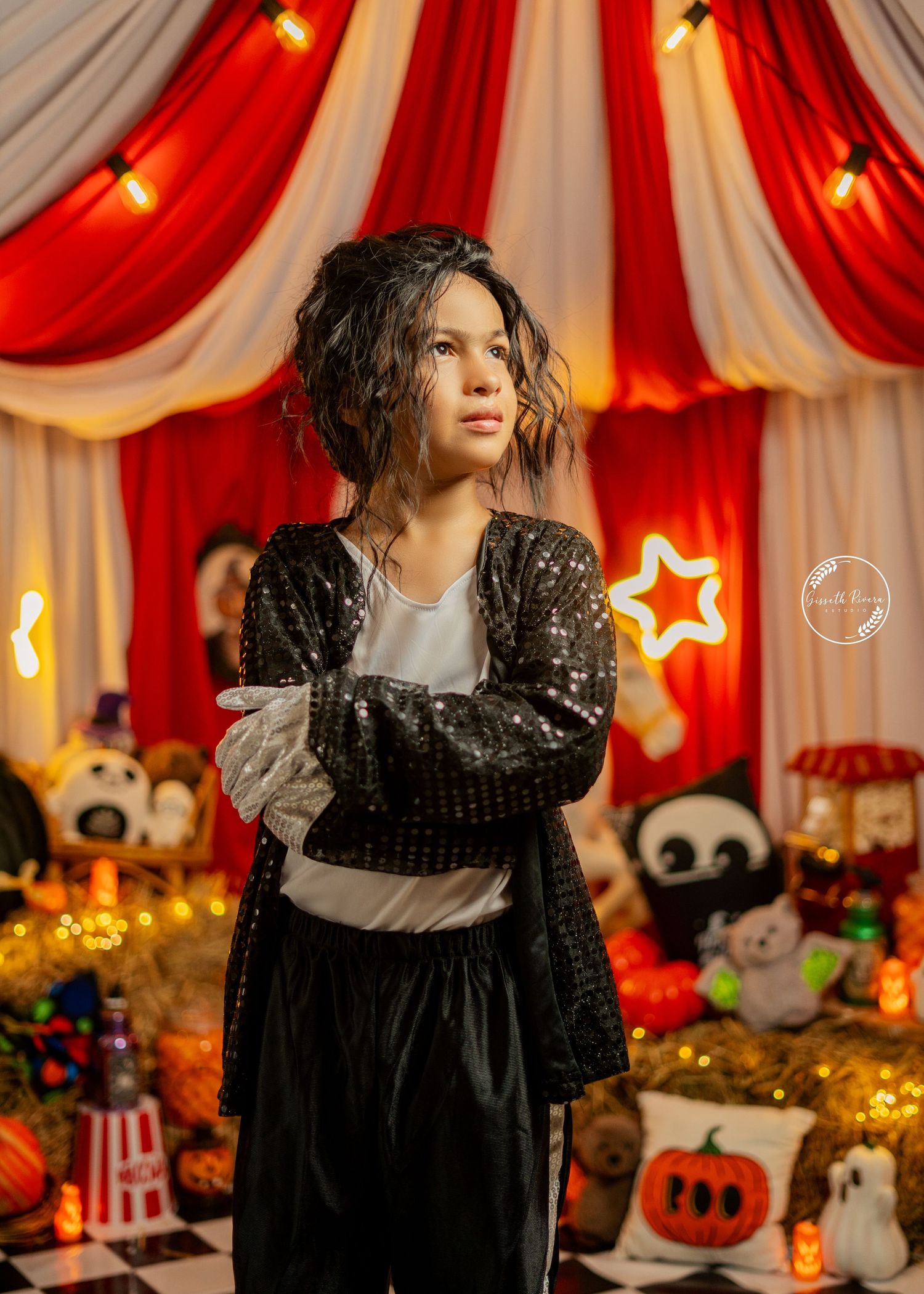 Child in sequin jacket poses at Halloween circus backdrop with plush toys and warm lights.