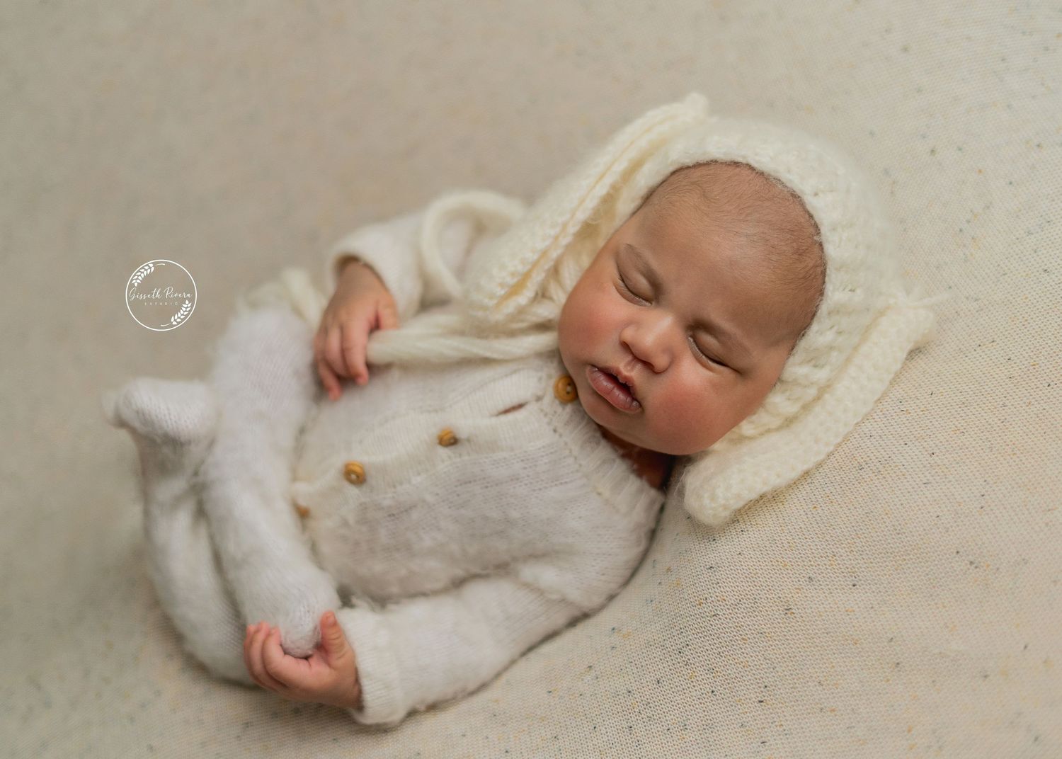 Sleeping newborn baby in cream knit outfit and bonnet rests peacefully on soft blanket.