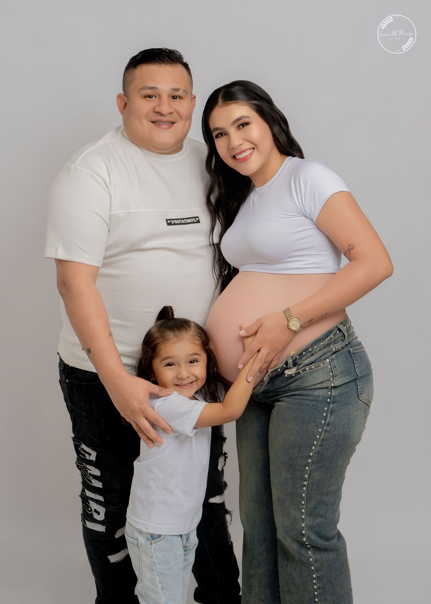Maternity family portrait with father, pregnant mother, and young daughter in white outfits on gray backdrop.
