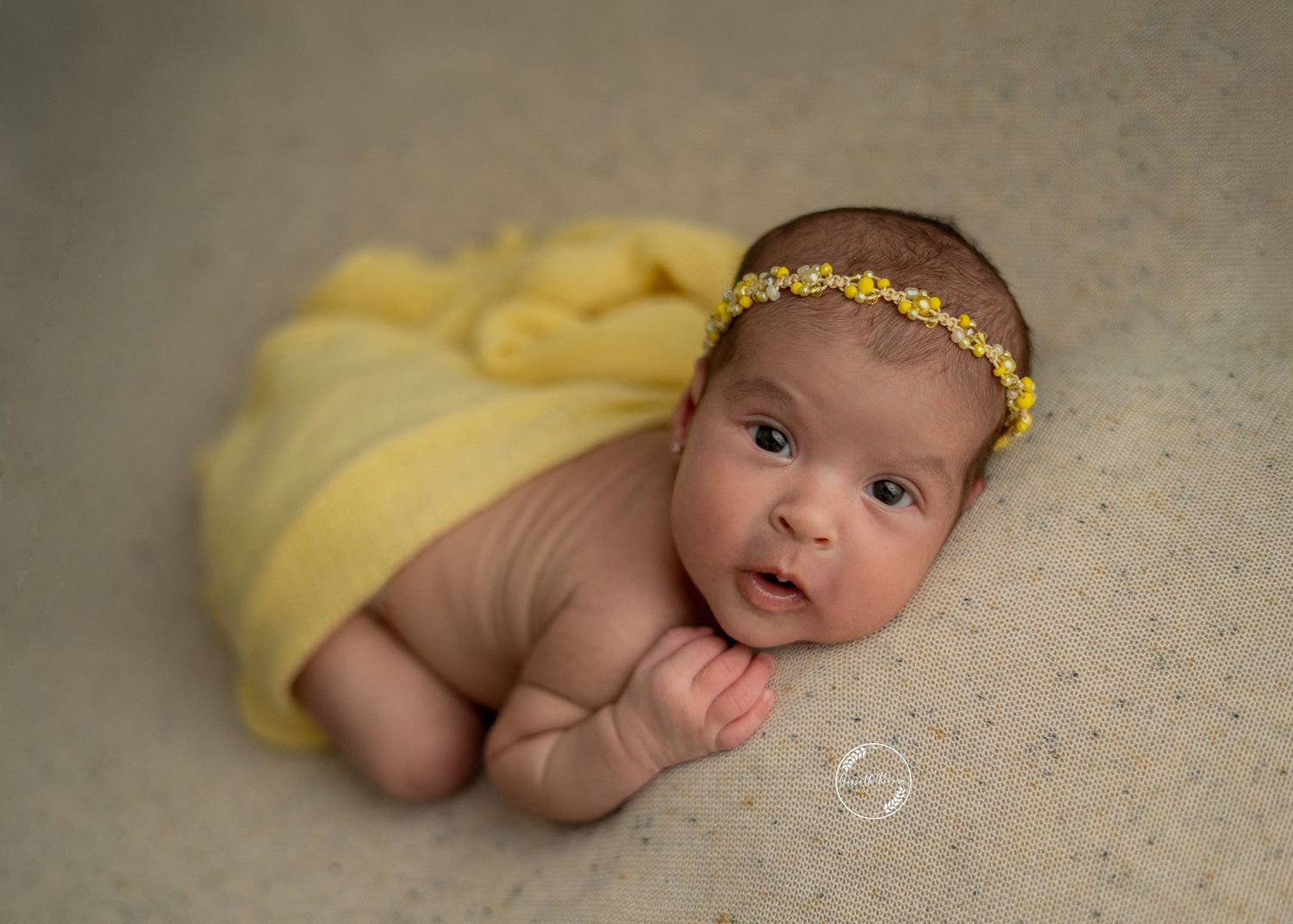 Alert newborn baby wrapped in yellow cloth with floral headband poses for studio portrait.