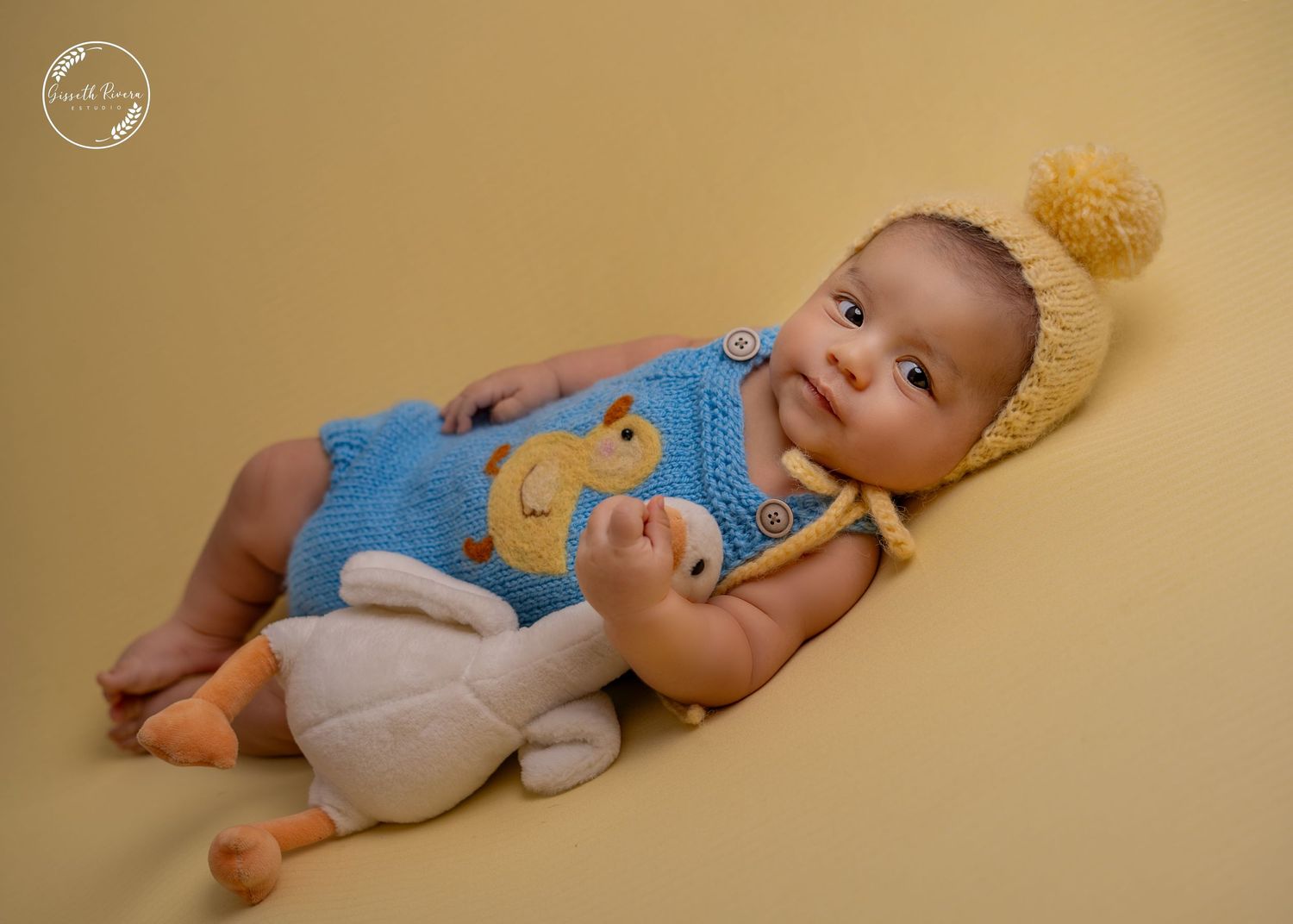 Adorable baby in blue knit bear romper and yellow pom-pom hat with stuffed toy.
