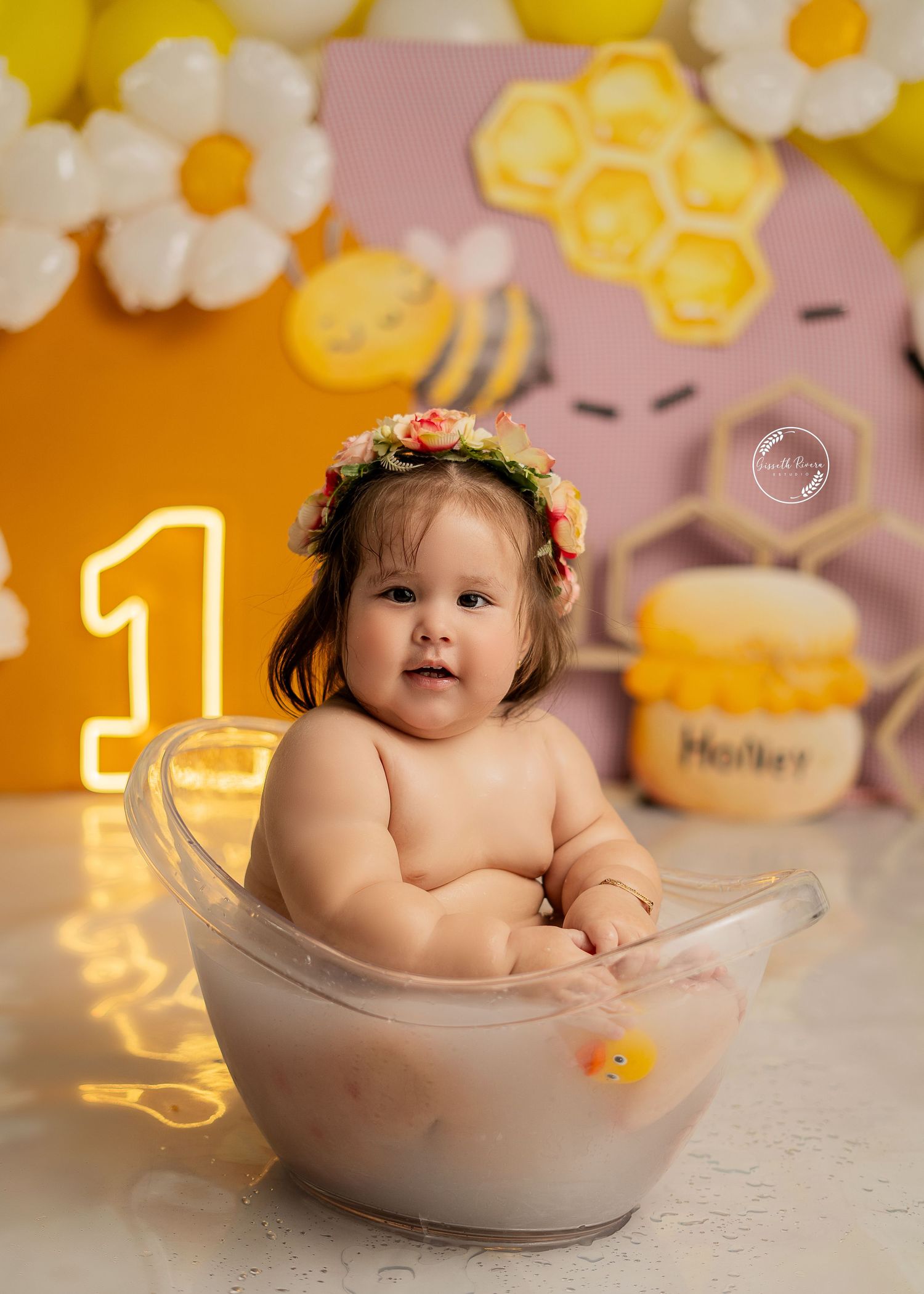 Baby girl in floral crown sits in clear tub during bee-themed first birthday bath smash.