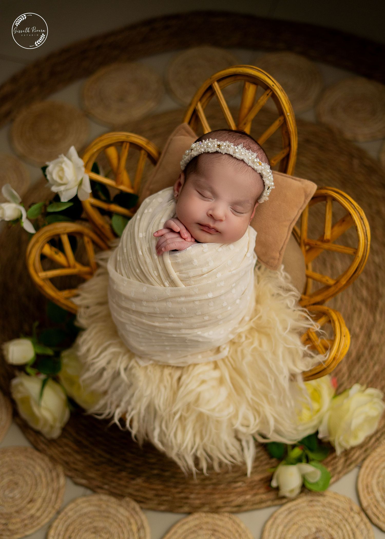 Swaddled newborn with pearl headband sleeps in decorative gold wagon with white roses.