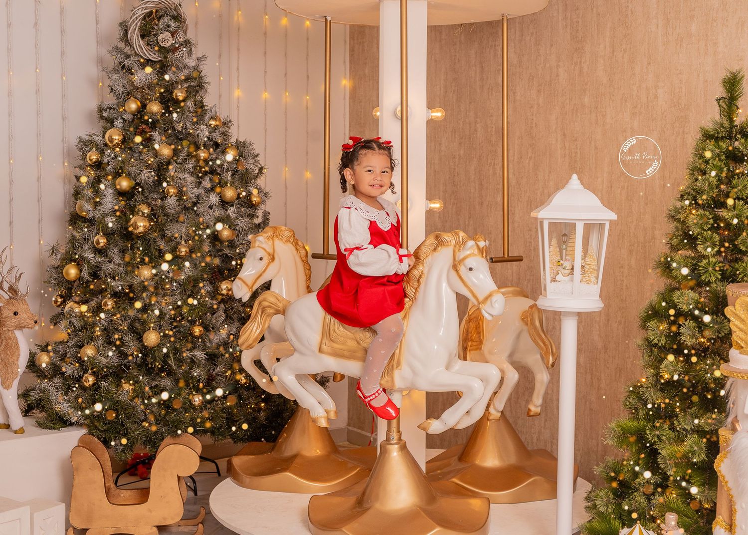 Young girl in red dress sits on carousel horses beside decorated Christmas trees in festive holiday photo session.