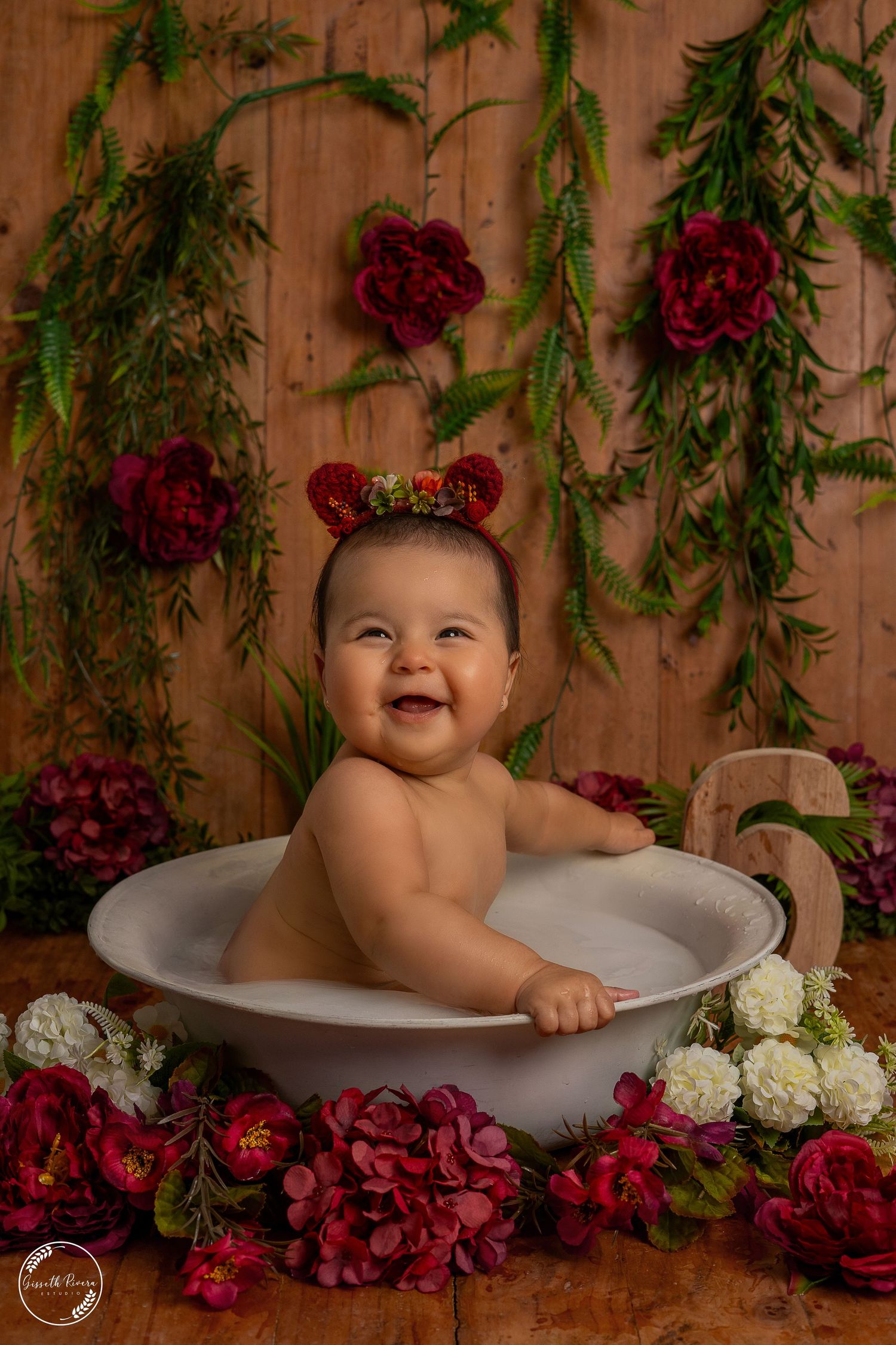 Joyful baby with floral headband sitting in a white tub, surrounded by red blooms on a wooden background.