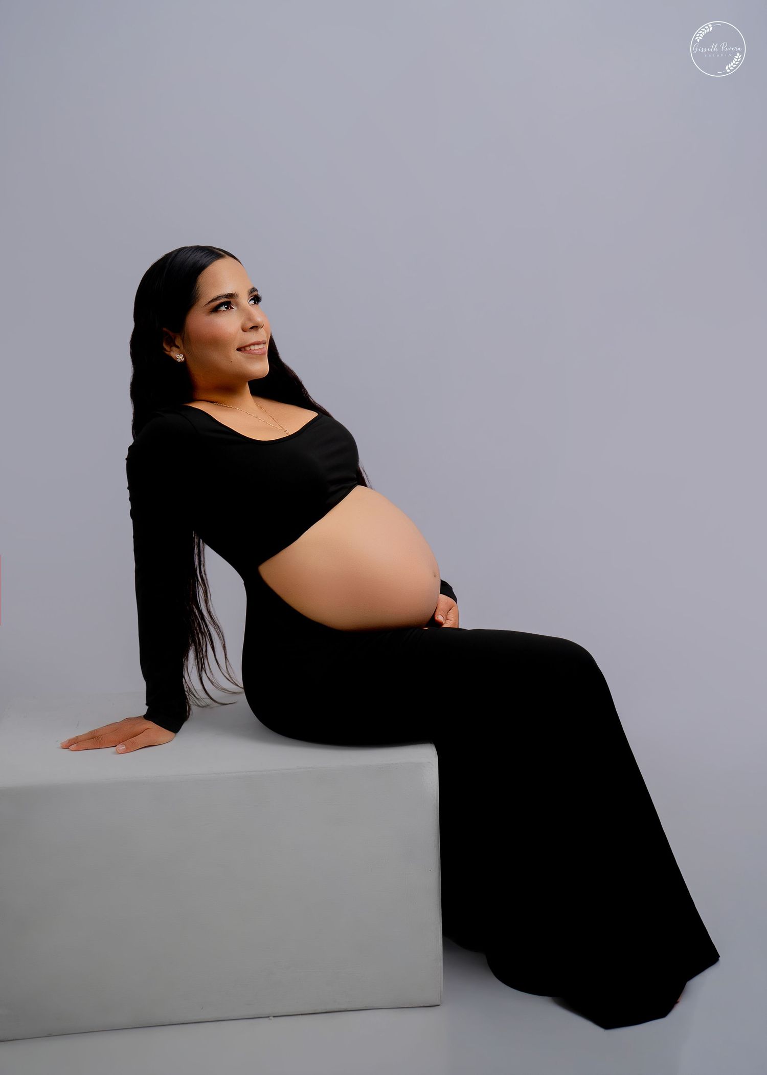 Elegant pregnant woman in black two-piece outfit seated on white cube against gray backdrop.