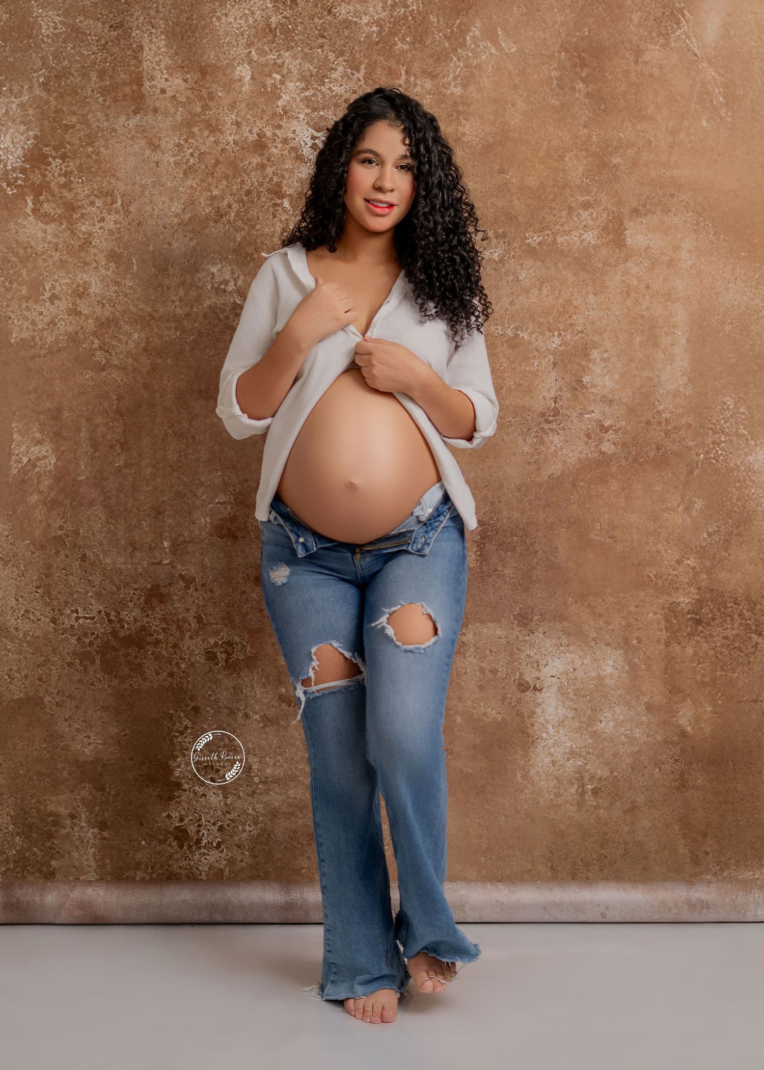 Pregnant woman with curly hair poses in white shirt and ripped jeans against textured backdrop.