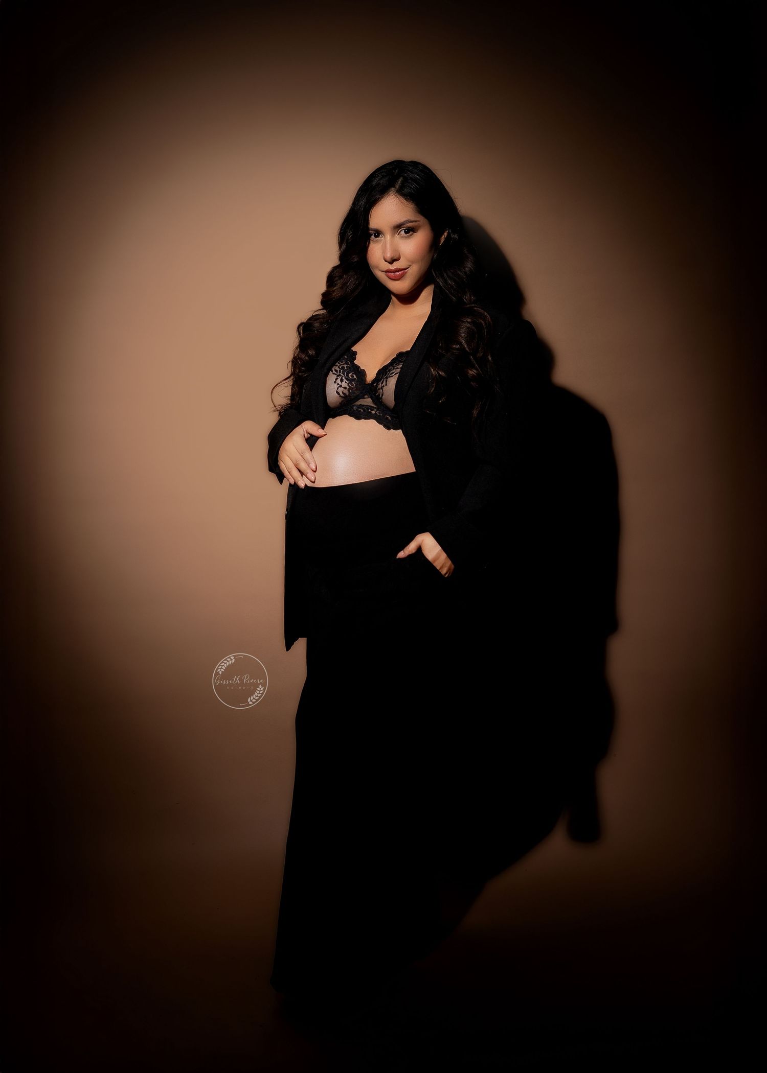 Pregnant woman in black lace bra and skirt poses elegantly against warm beige studio backdrop.