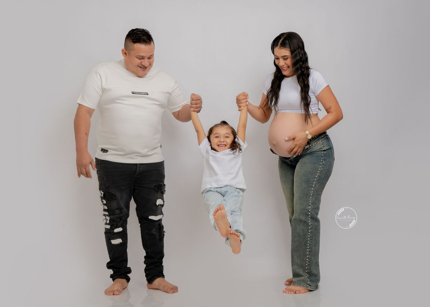 Maternity photo of parents lifting toddler by hands with pregnant mother on gray backdrop.