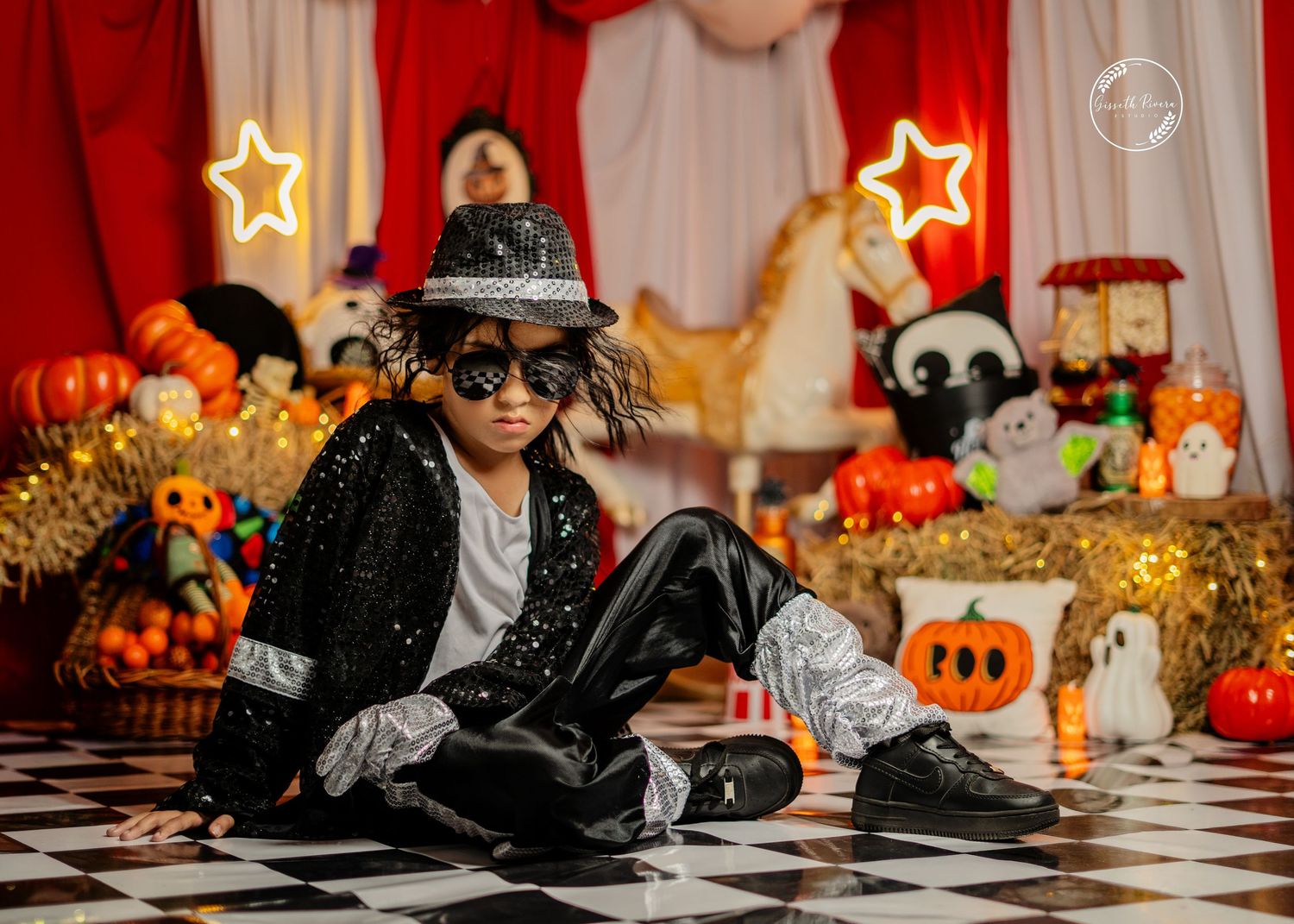 Child in Michael Jackson outfit with sunglasses and fedora sits on checkered floor with Halloween decor.