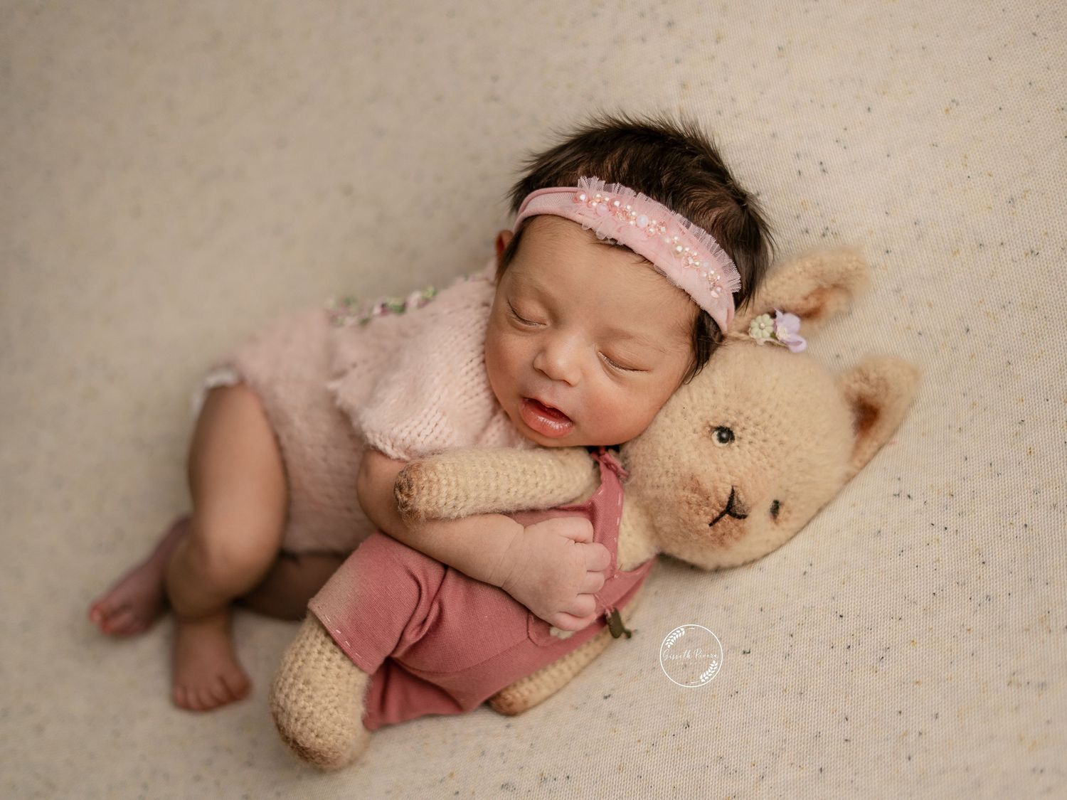 Sleeping newborn in pink outfit cuddles crocheted teddy bear in professional studio photo.