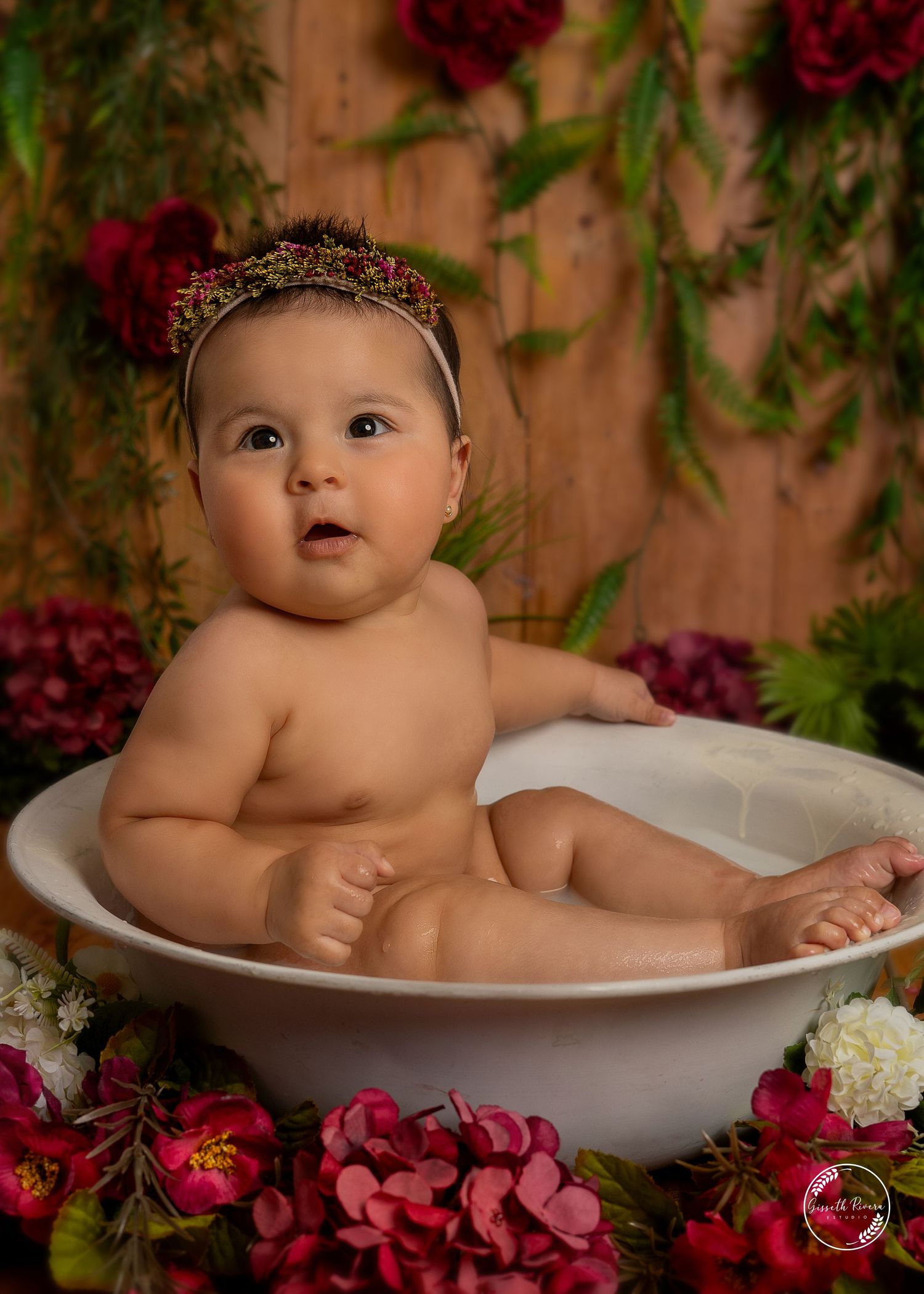 Adorable baby girl with floral crown sits in white bowl bath surrounded by red and pink flowers.