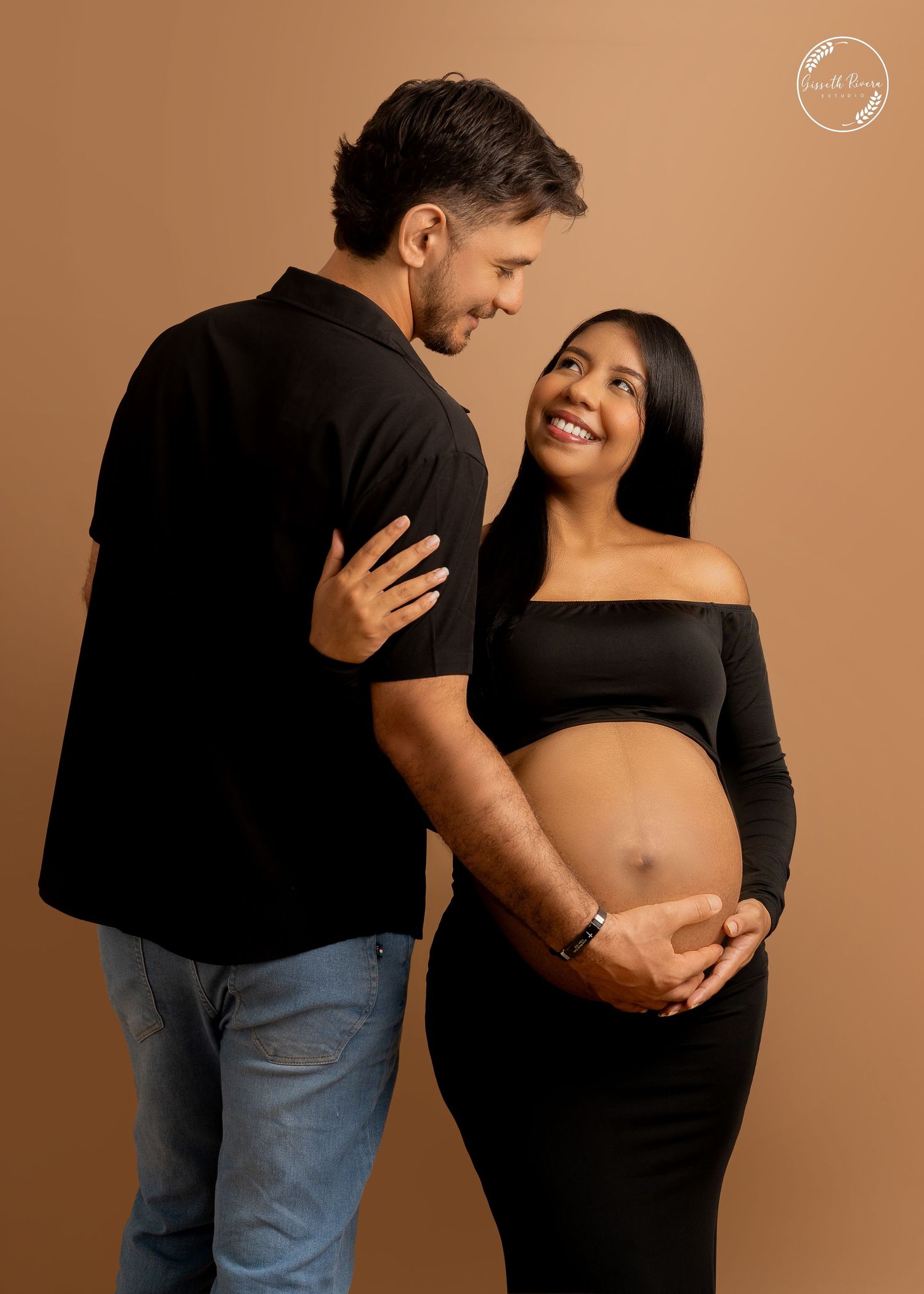 Couple maternity portrait in matching black outfits with man embracing pregnant woman on brown backdrop.