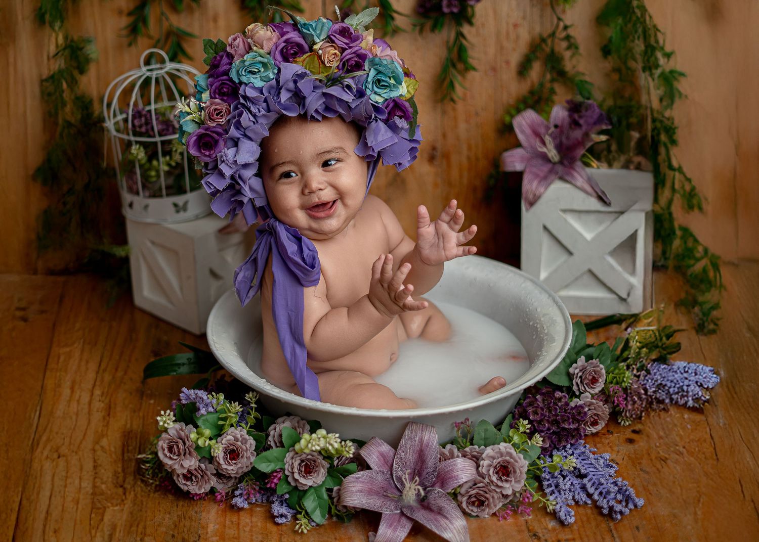 Happy baby in a milk bath tub wearing a purple floral bonnet, surrounded by colorful flowers and decor.