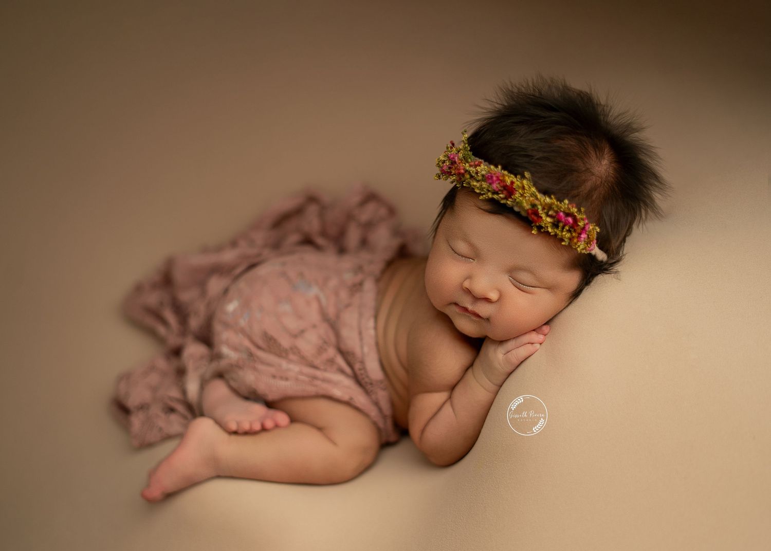 Sleeping newborn wrapped in mauve cloth with floral headband rests on beige background.