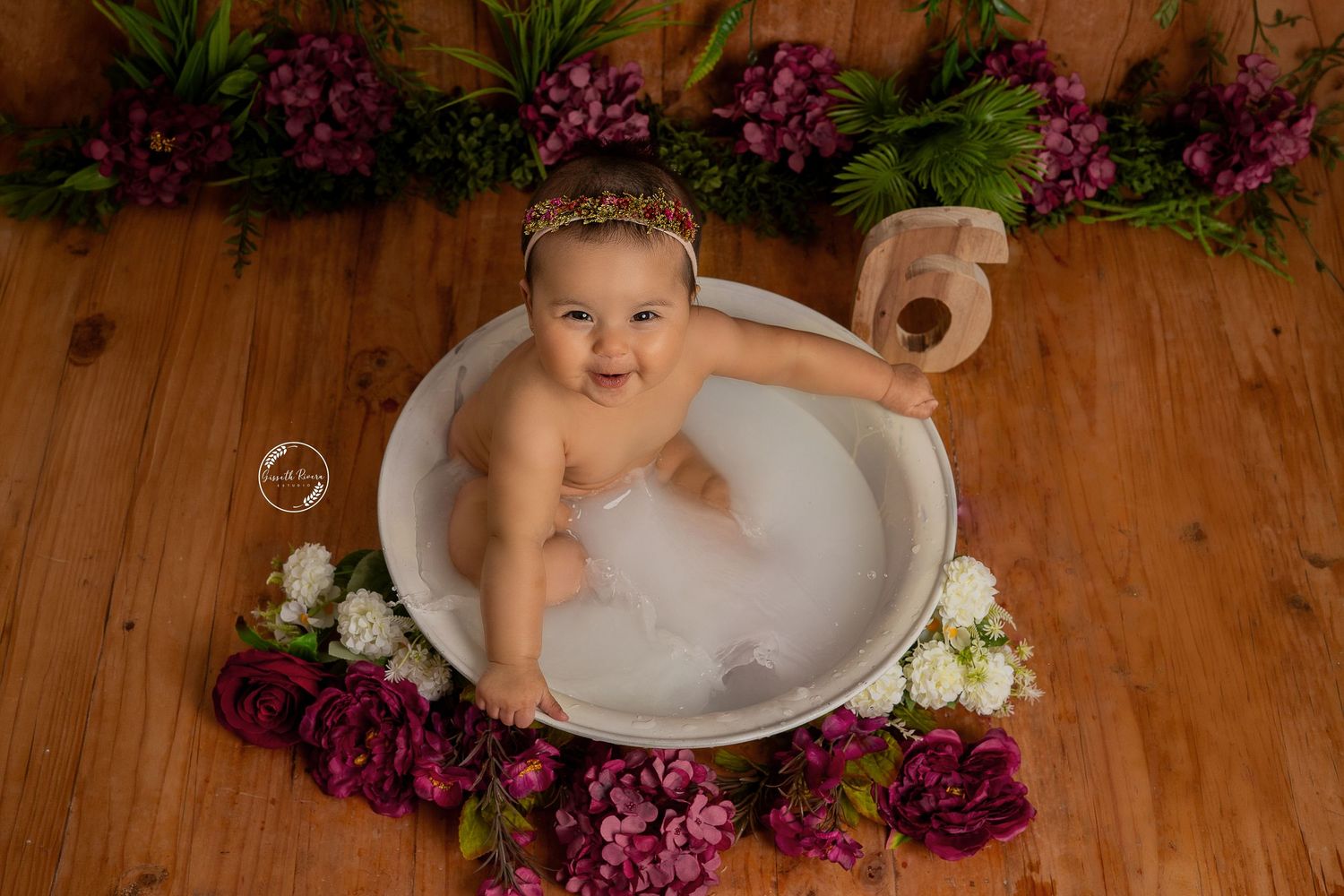 Smiling baby sitting in a white tub with bubbles, surrounded by red and white flowers on a wooden floor.