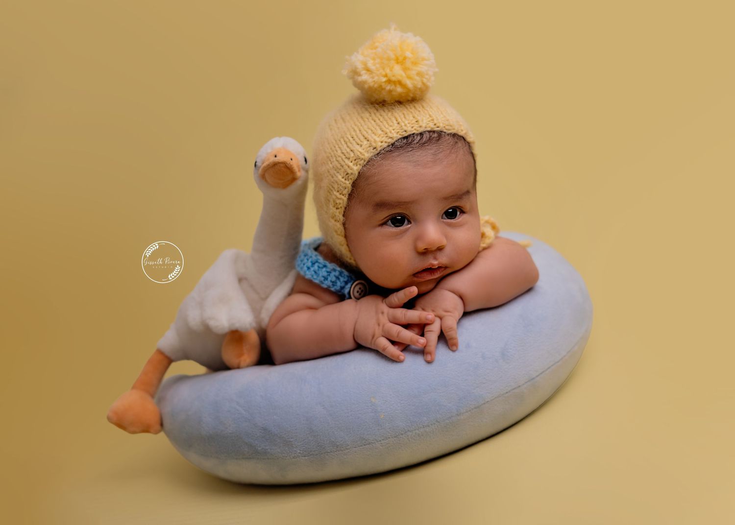 Cute baby in yellow knit hat resting on blue donut pillow with duck plush toy.