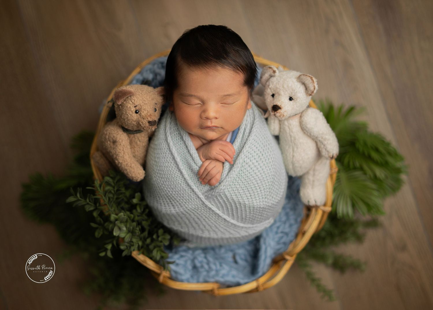 Peacefully sleeping newborn wrapped in blue swaddle in wicker basket with teddy bears and green ferns.