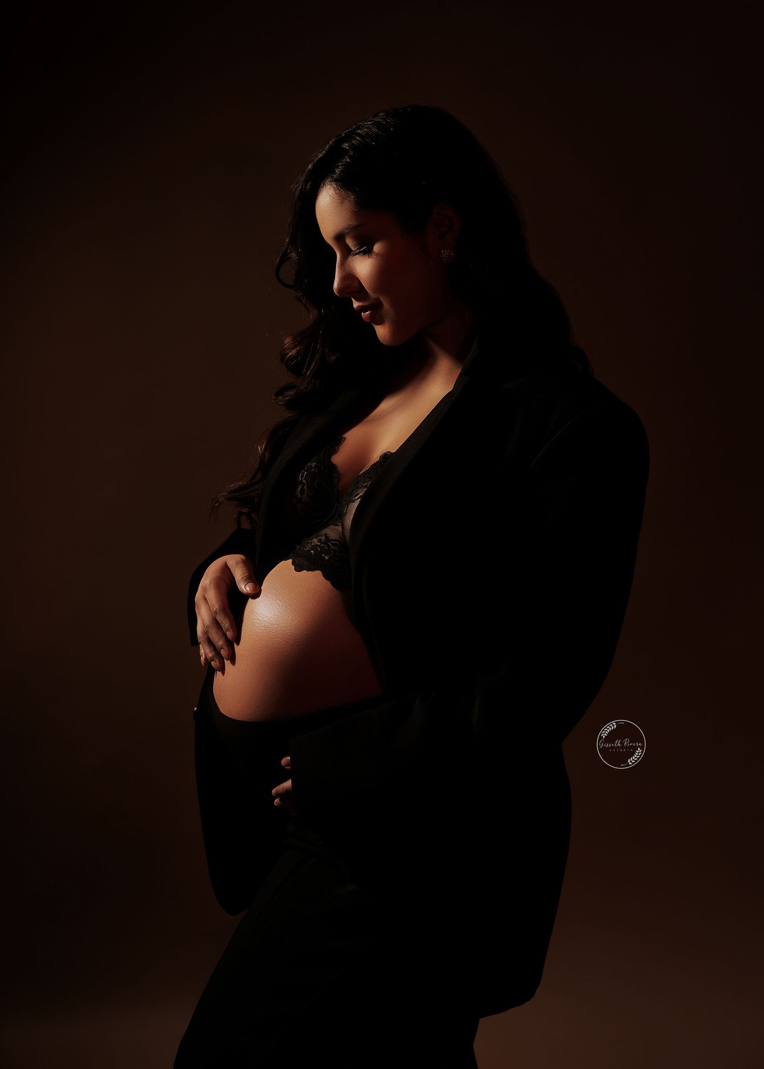 Pregnant woman in black blazer cradling baby bump in dramatic dark studio lighting.