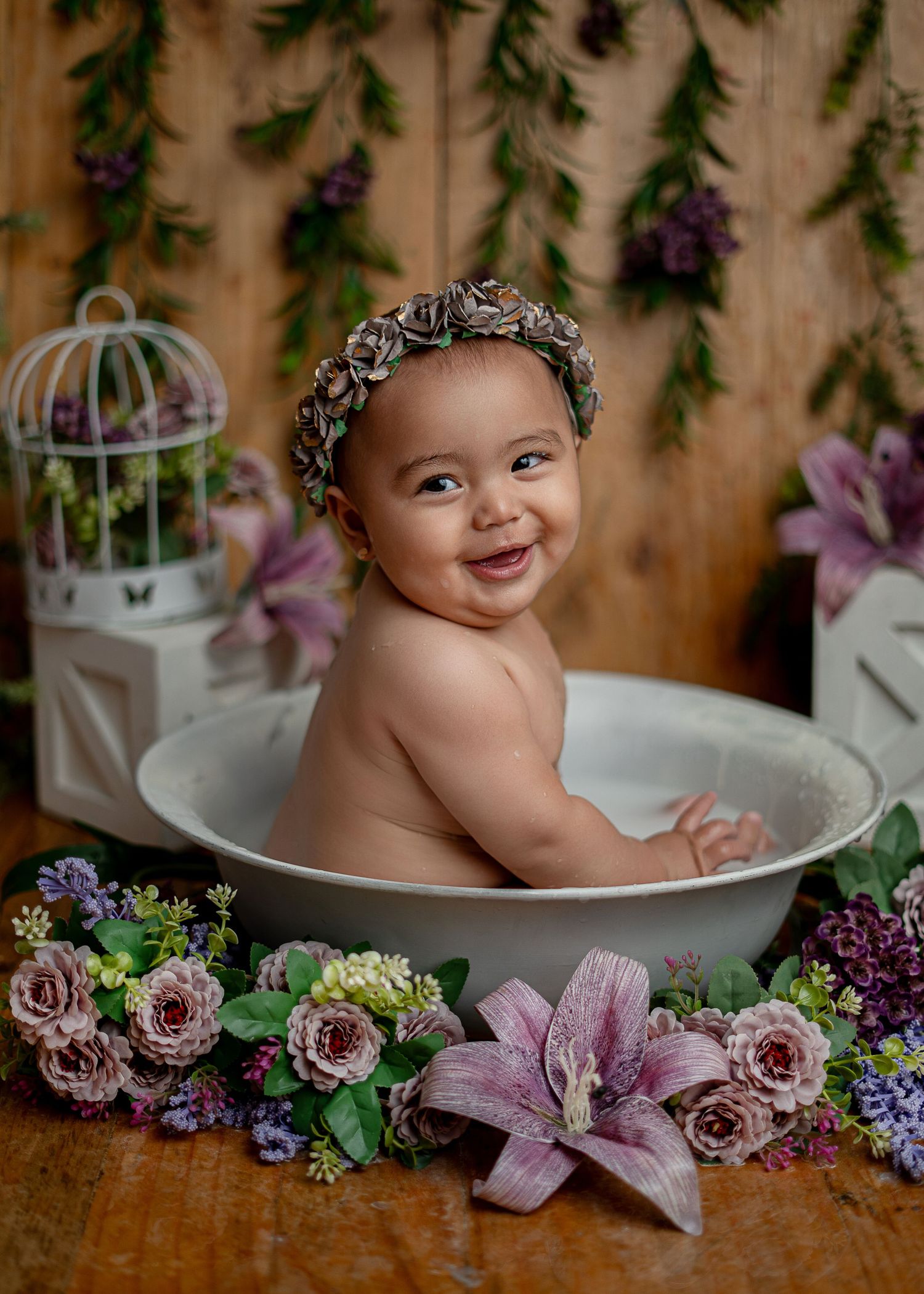 Smiling baby wearing a floral headband sitting in a white tub with pink lilies and purple flowers nearby.